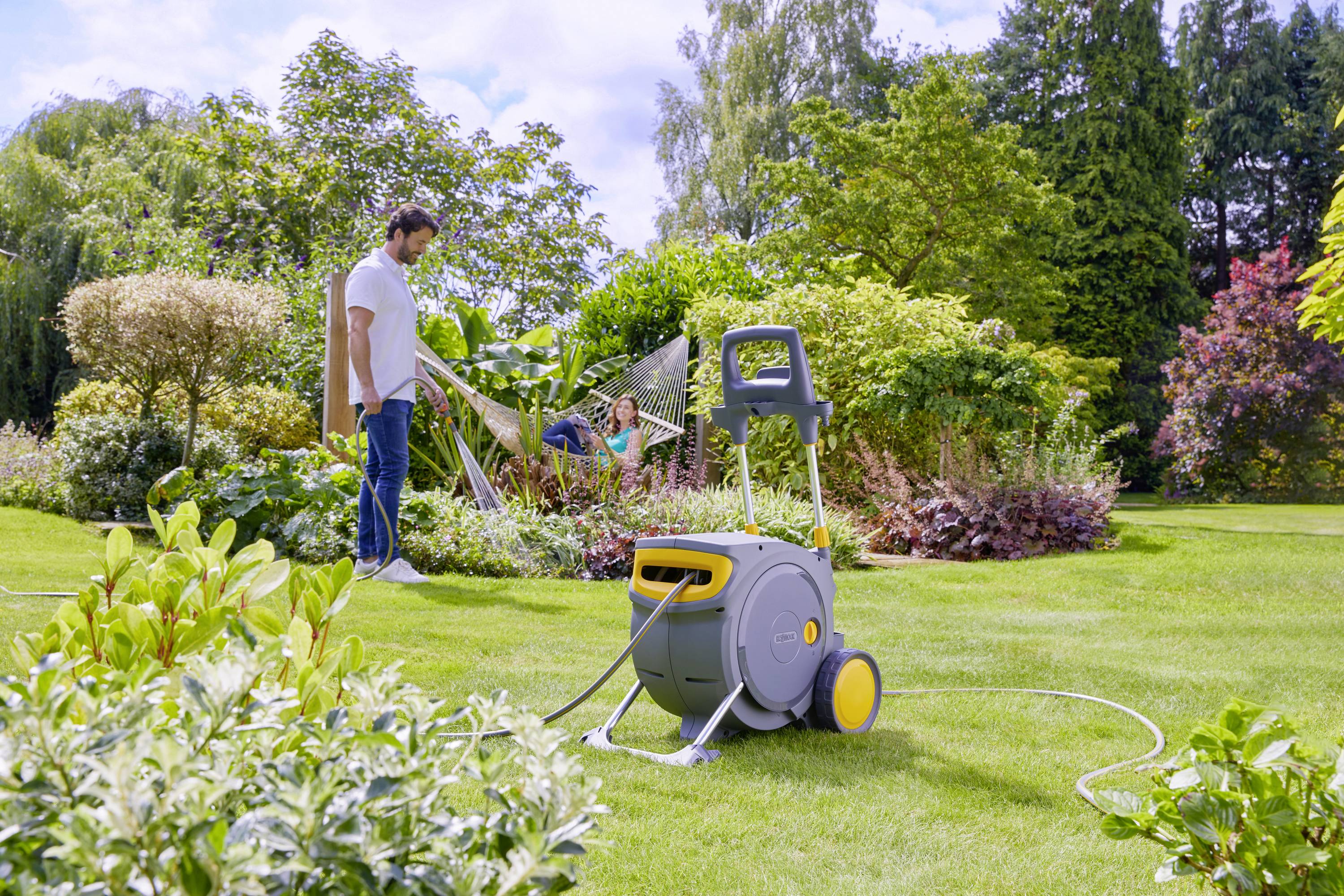 Ein Mann gießt Pflanzen im Garten mit einem Wasserschlauch. Eine Person liegt in einer Hängematte und entspannt sich.