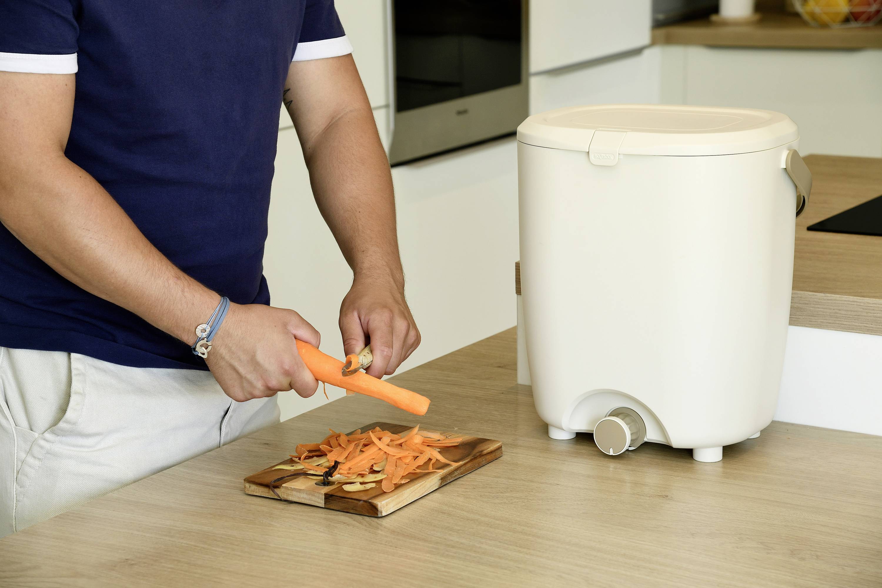 Eine Person schält eine Karotte auf einem Schneidebrett in der Küche. Ein weißer Komposteimer steht auf der Arbeitsplatte.
