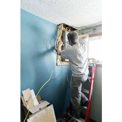 Ein Handwerker auf einer Leiter repariert elektrische Kabel in einer Wand. Die Wand ist blau, es gibt ein offenes Fenster daneben.
