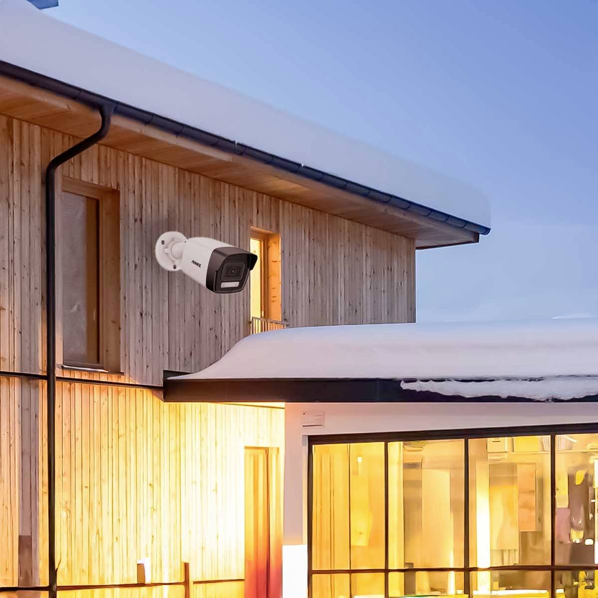 Holzhaus mit Schnee auf dem Dach, überwacht von einer außen angebrachten Sicherheitskamera, beleuchtet von warmer Innenbeleuchtung.
