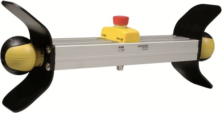 Ein Sicherheitsschalter mit einem gelben Gehäuse und rotem Knopf, montiert auf einer Metallstange mit schwarzen Schutzabdeckungen.