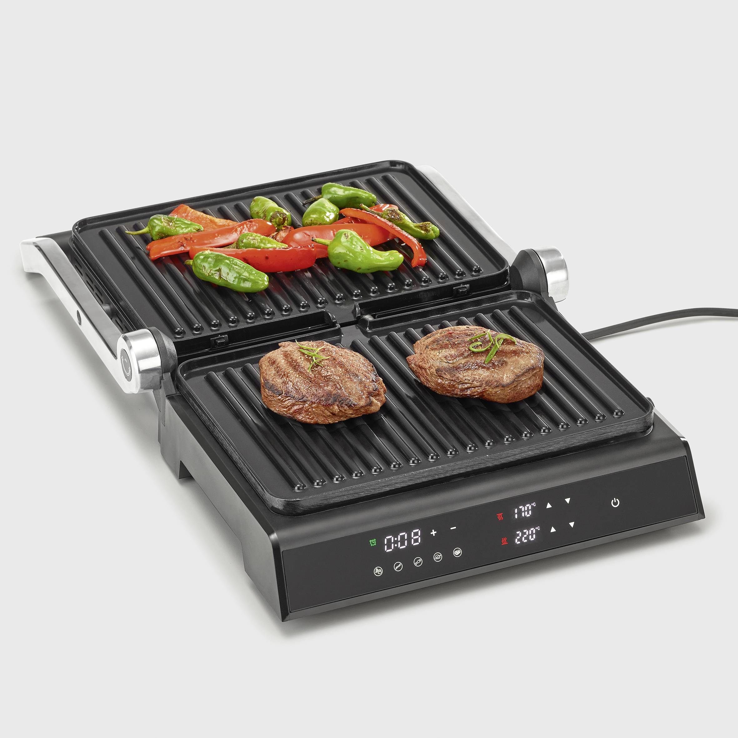 Ein elektrischer Kontaktgrill mit zwei Steaks auf der unteren Platte und gegrilltem Gemüse auf der oberen. Temperaturanzeige ist sichtbar.