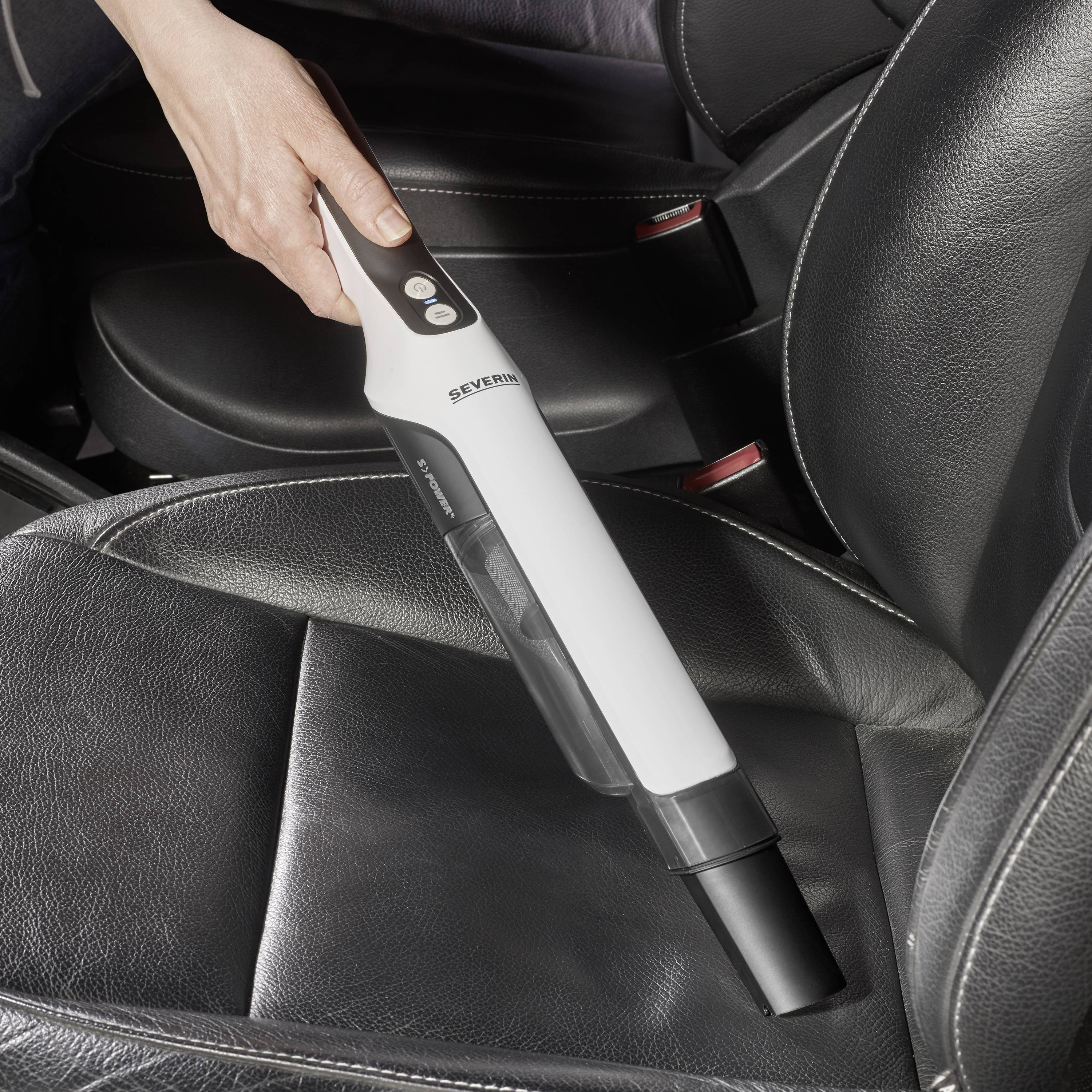 Eine Person reinigt einen Autositz mit einem weißen Handstaubsauger. Der Sitz ist aus schwarzem Leder.