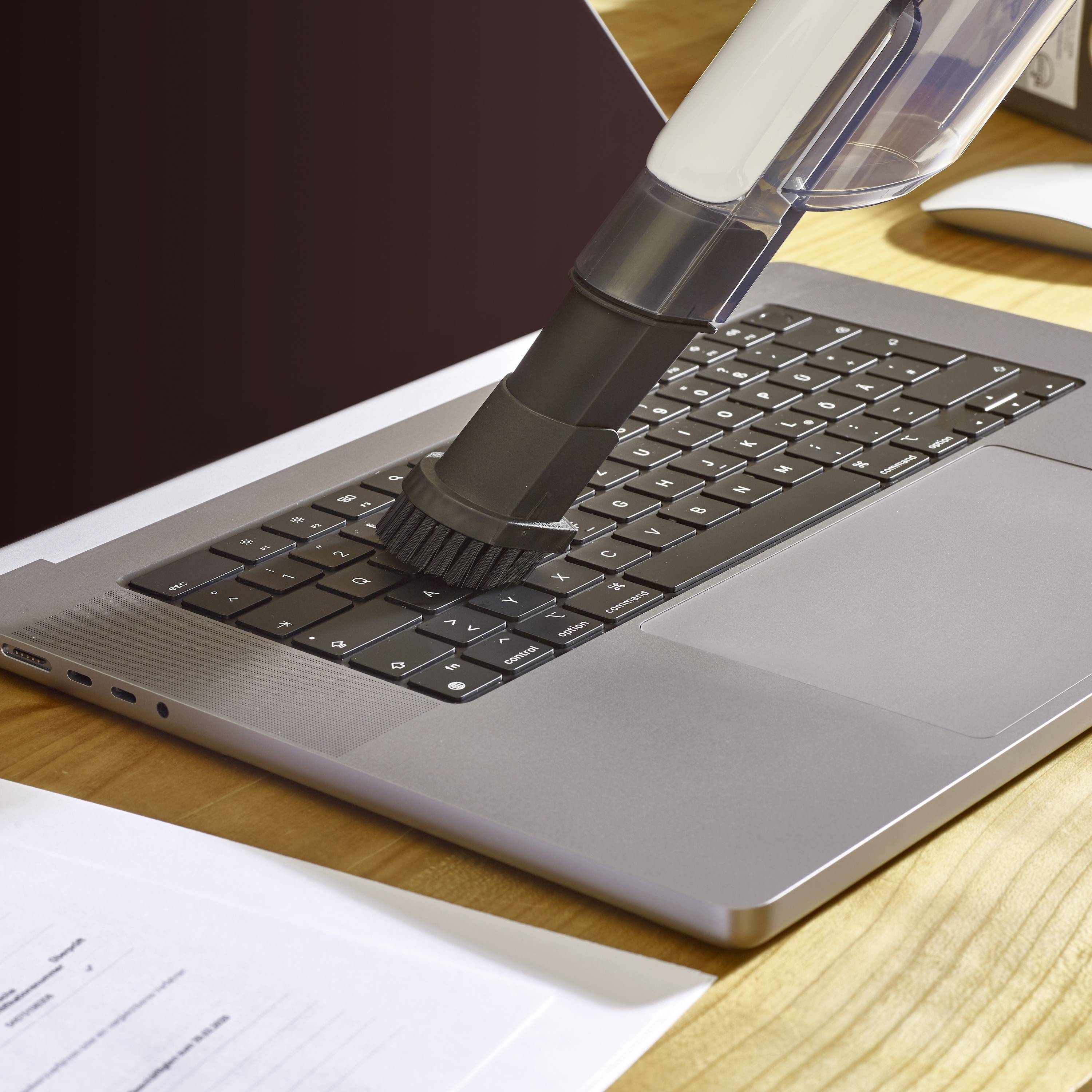 'Ein Staubsaugeraufsatz reinigt die Tastatur eines Laptops auf einem Schreibtisch aus Holz, neben Papieren und einem Computermaus.'