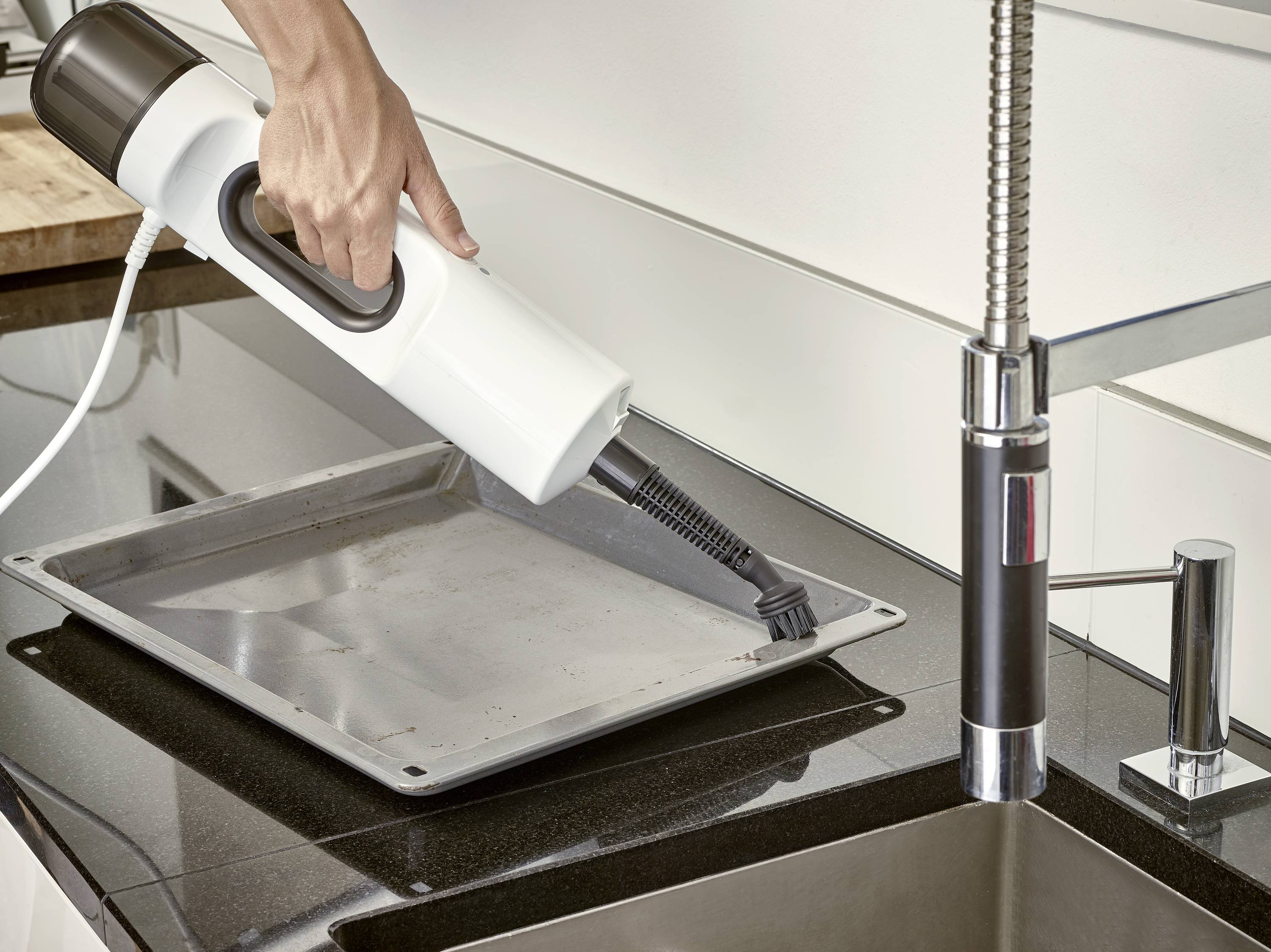 Eine Hand reinigt ein Backblech auf einer Küchenarbeitsplatte mit einem Dampfreiniger. Daneben befindet sich ein Wasserhahn.