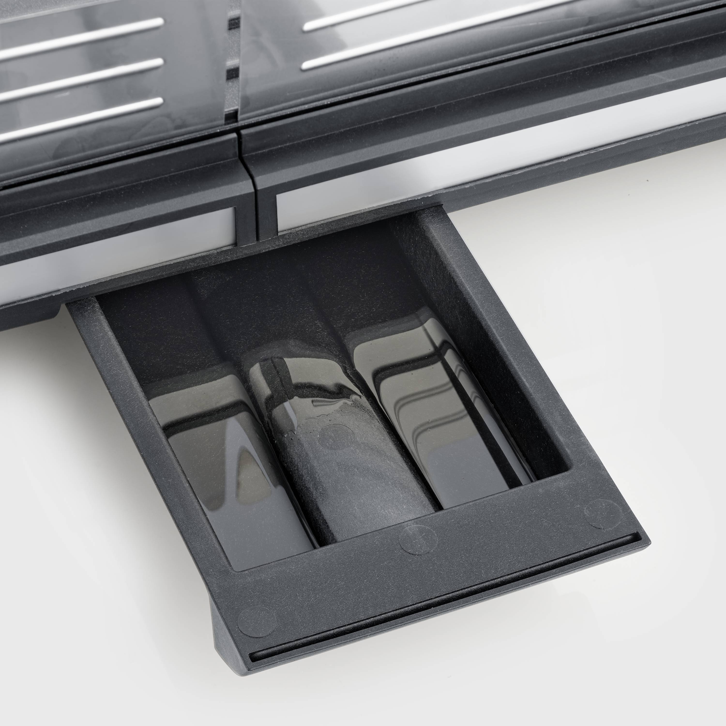 Ein schwarzer, herausgezogener Ablagebehälter unter einem grauen Gerät, möglicherweise ein Toaster. Der Behälter scheint abnehmbar zu sein.