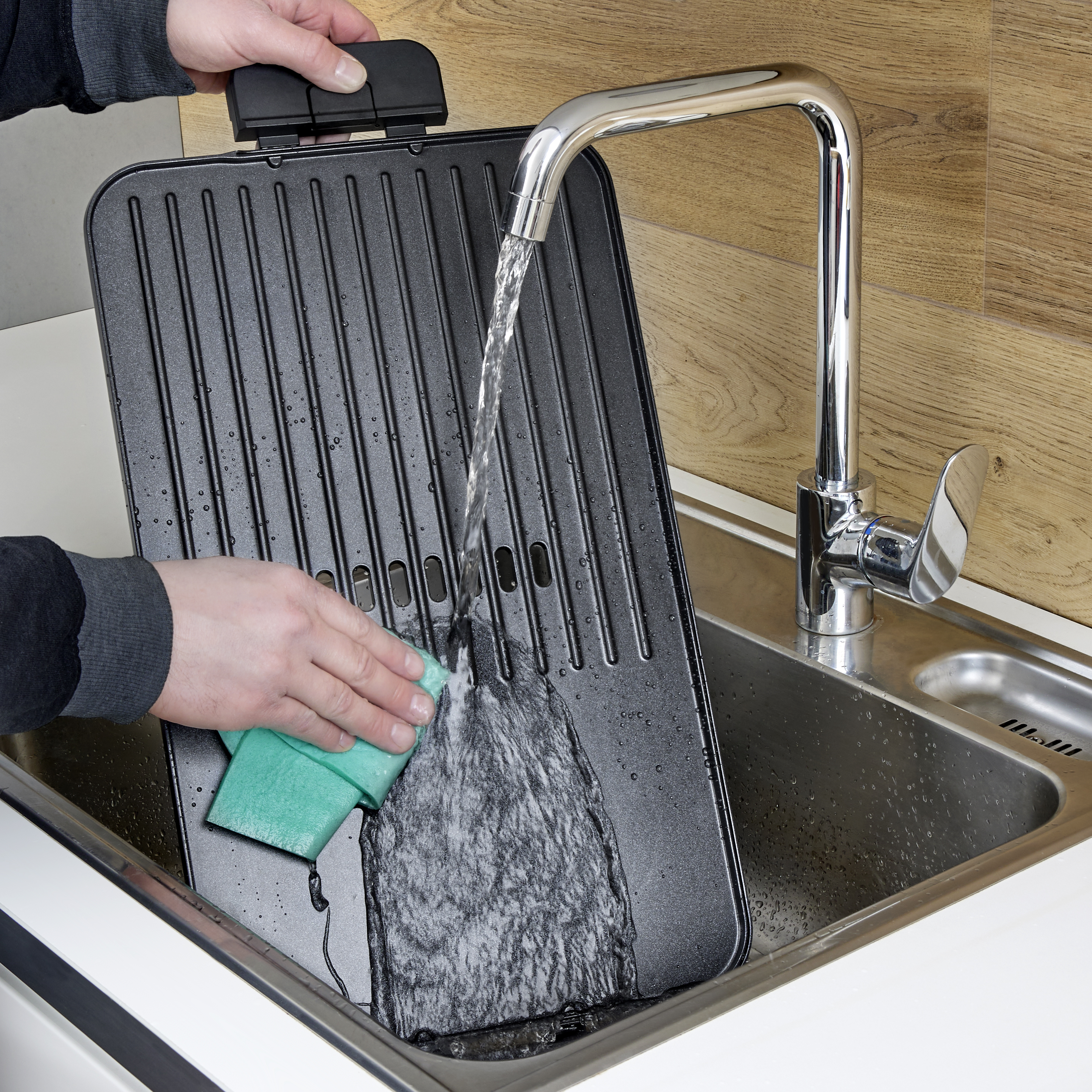 Eine Person reinigt ein Waffeleisen mit einem grünen Schwamm unter fließendem Wasser in einem Spülbecken.