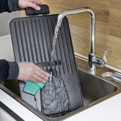 Eine Person reinigt ein Waffeleisen mit einem grünen Schwamm unter fließendem Wasser in einem Spülbecken.