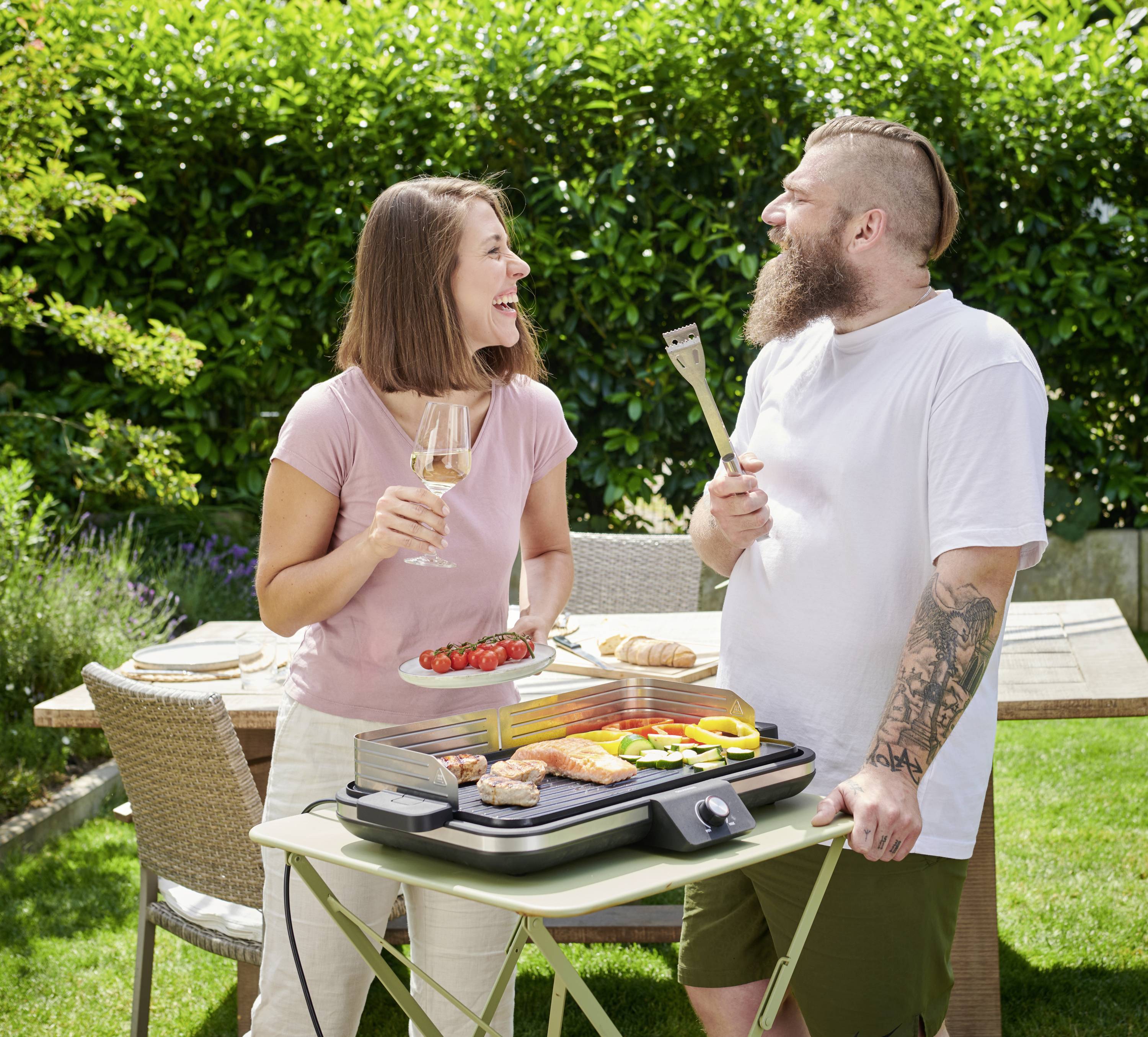 Zwei Personen lächeln sich an, während sie im Garten grillen. Sie halten einen Teller mit Essen und Grillzubehör. Im Hintergrund ein Tisch mit Geschirr.