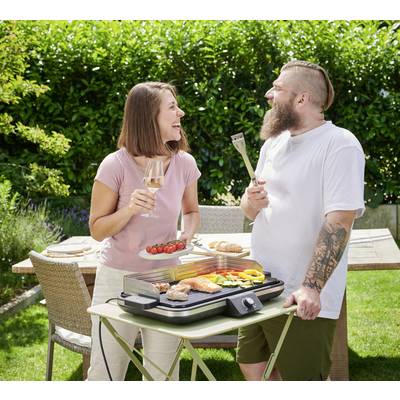 Zwei Personen lächeln sich an, während sie im Garten grillen. Sie halten einen Teller mit Essen und Grillzubehör. Im Hintergrund ein Tisch mit Geschirr.