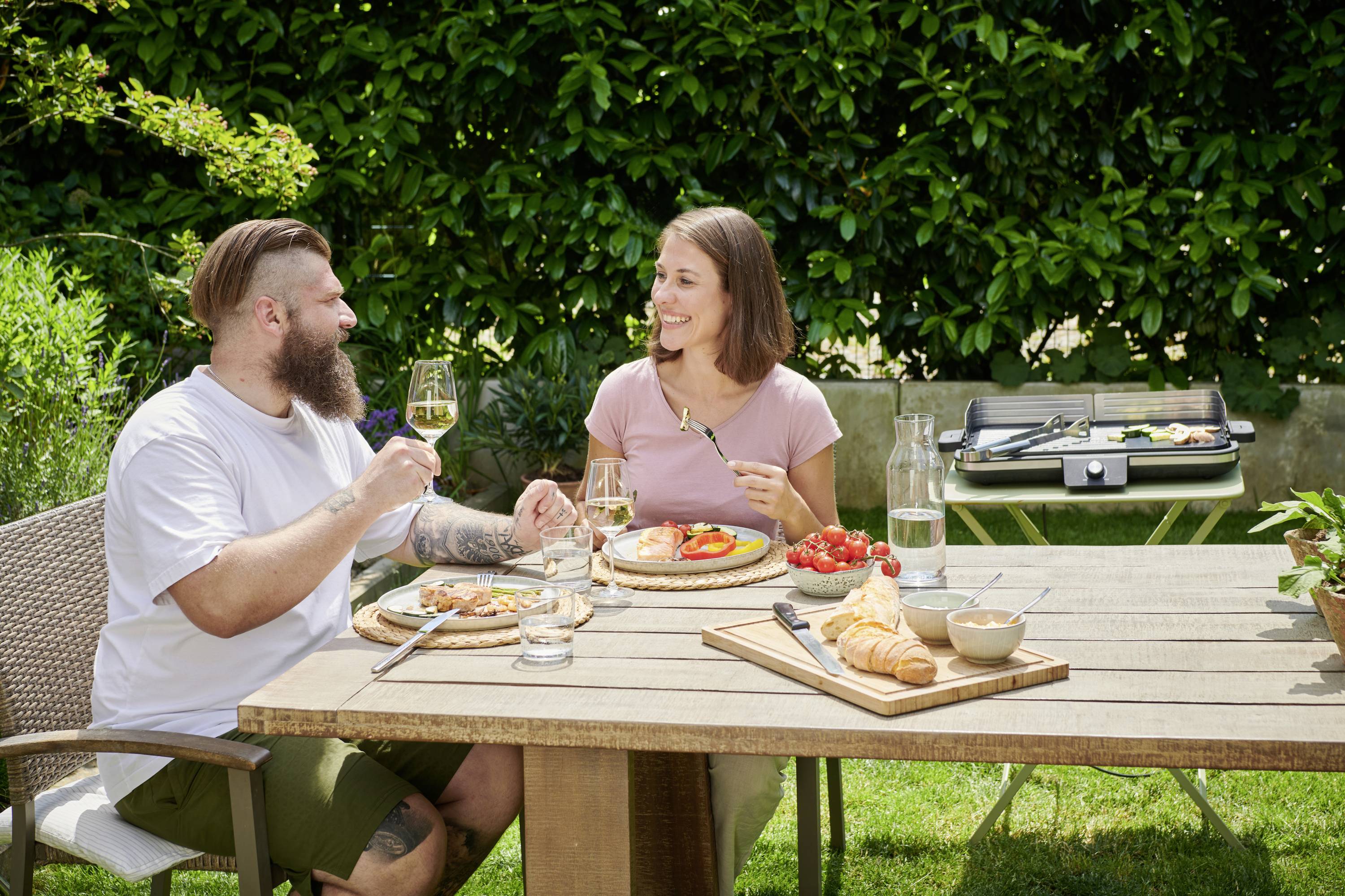 Ein Mann und eine Frau sitzen an einem Tisch im Garten und essen. Sie lächeln und heben ihre Gläser zum Anstoßen. Im Hintergrund ist ein Grill zu sehen.