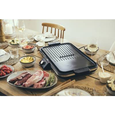 Ein Tischgrill steht in der Mitte eines gedeckten Tisches mit verschiedenen Schalen voller Zutaten wie Fleisch, Tomaten und Ananas.