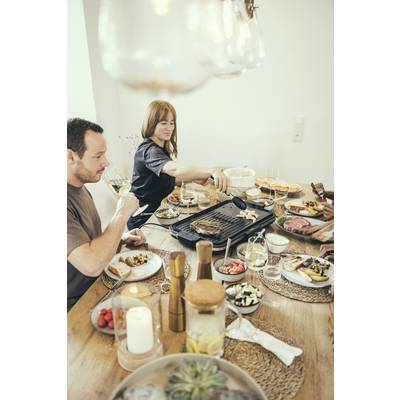 Ein Mann und eine Frau sitzen an einem gedeckten Tisch beim Essen. Die Frau bedient eine Grillplatte, während der Mann ein Glas Wein hält.