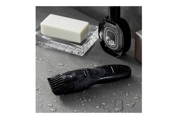 Ein nasser, schwarzer elektrischer Trimmer liegt auf einer grauen Oberfläche neben einer weißen Seife und einer Parfümflasche.