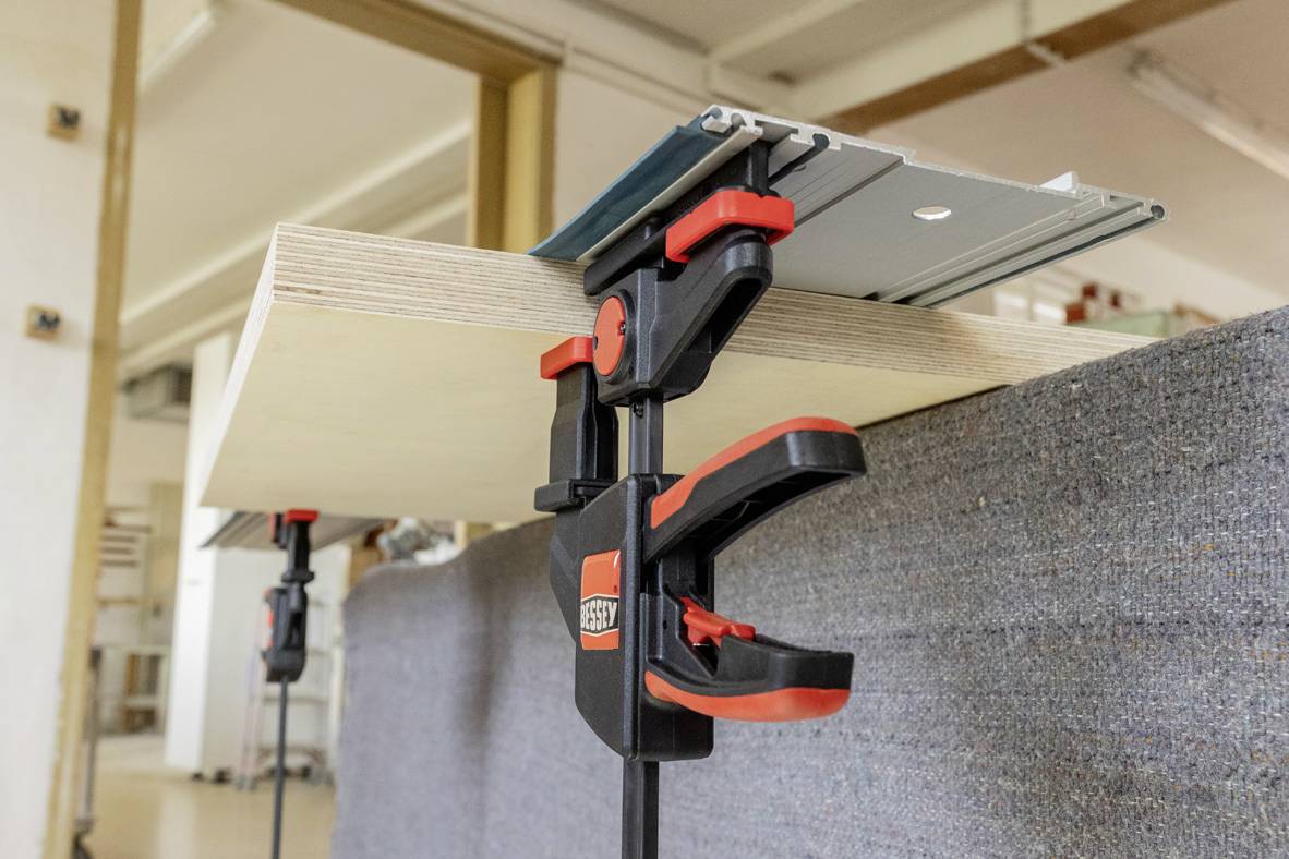 Ein Holzbrett wird mit zwei großen Schraubzwingen an einem Tisch befestigt. Im Hintergrund ist eine Werkstattumgebung sichtbar.