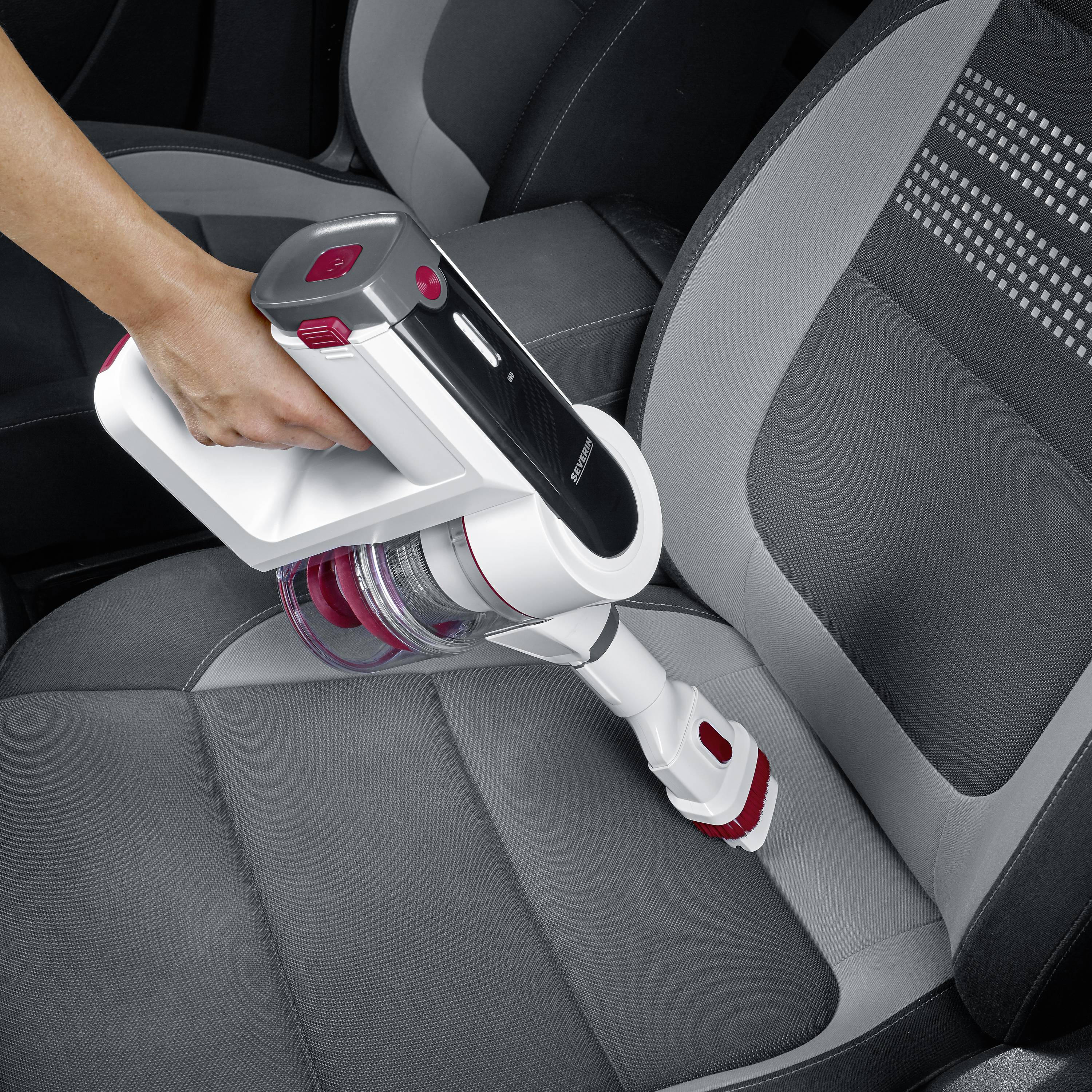 Eine Person reinigt den Autositz mit einem kabellosen Handstaubsauger. Der Sitz ist grau mit schwarzen Akzenten.