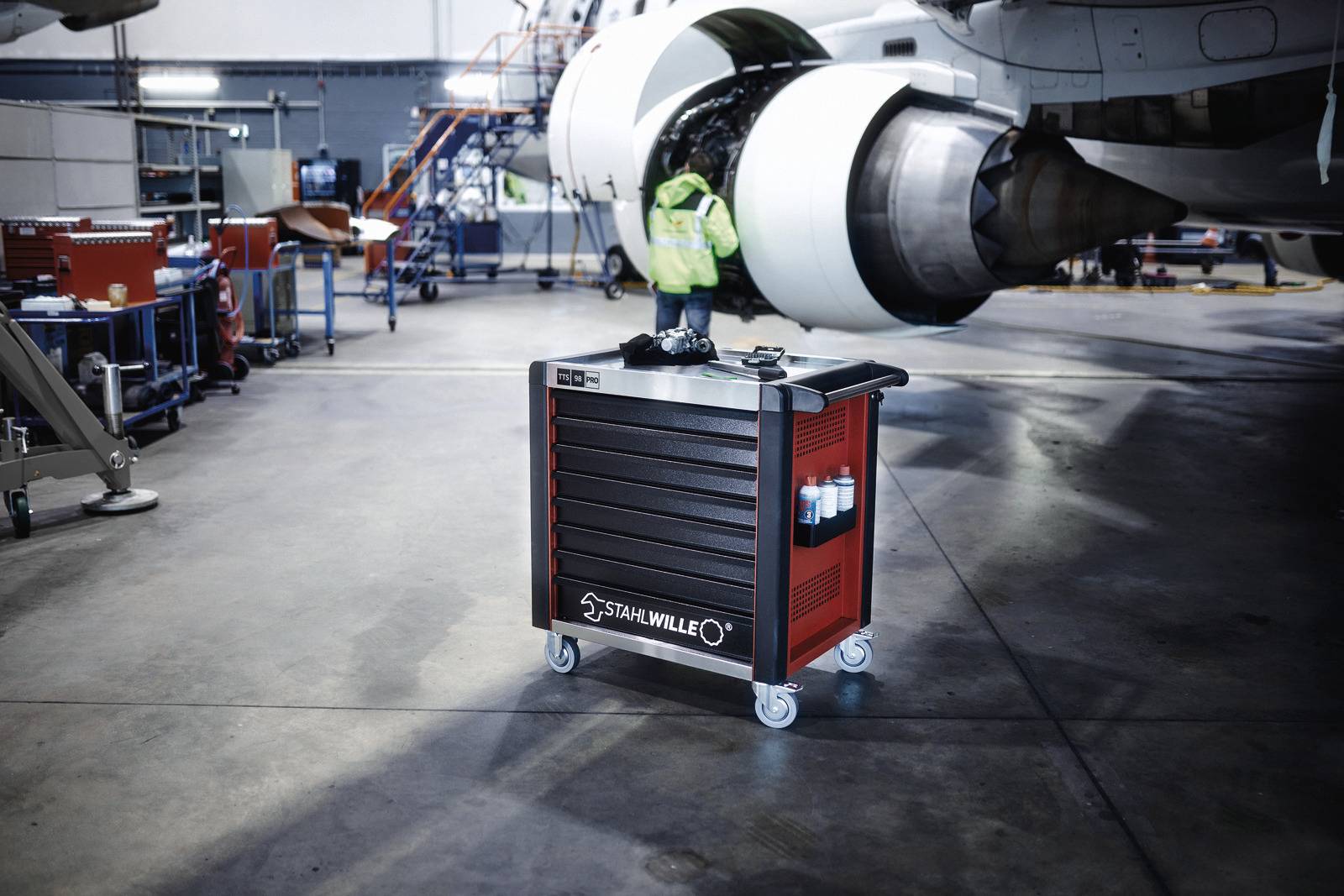 Ein roter Werkzeugwagen mit verschiedenen Fächern steht in einem Flugzeughangar, in dem ein Flugzeug zur Wartung bereitsteht.
