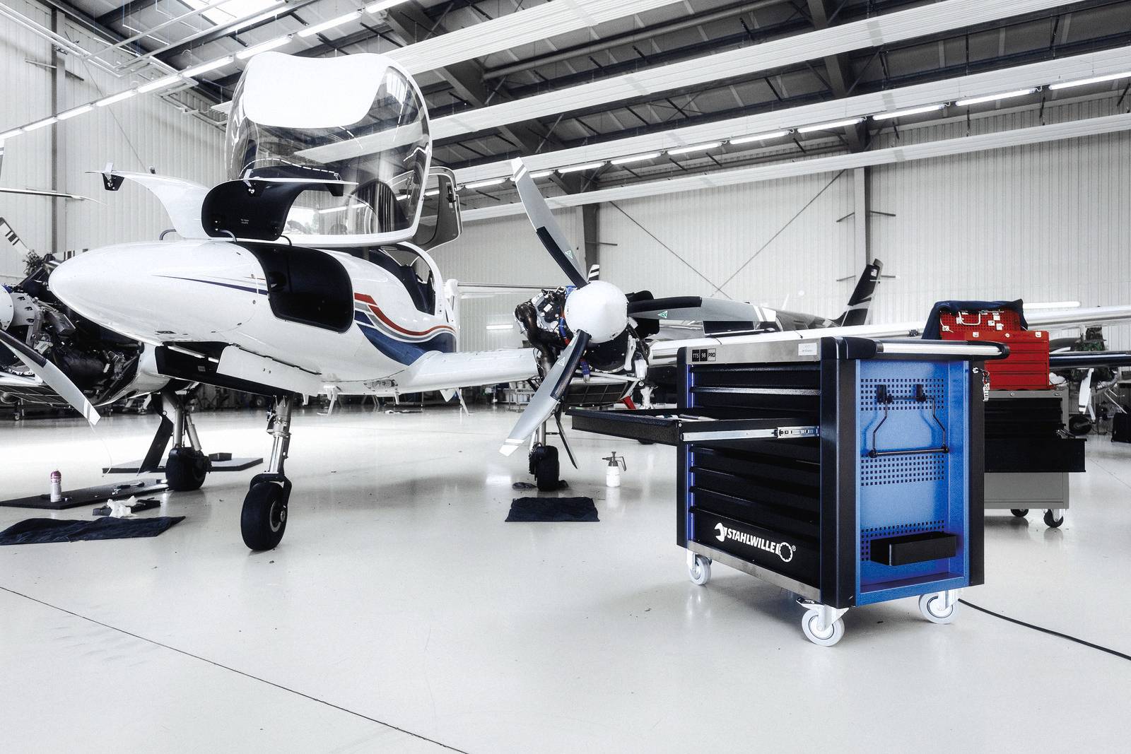 Ein kleines Flugzeug steht in einem Hangar. Daneben befindet sich ein blauer Werkzeugwagen. Die Umgebung ist hell und aufgeräumt.