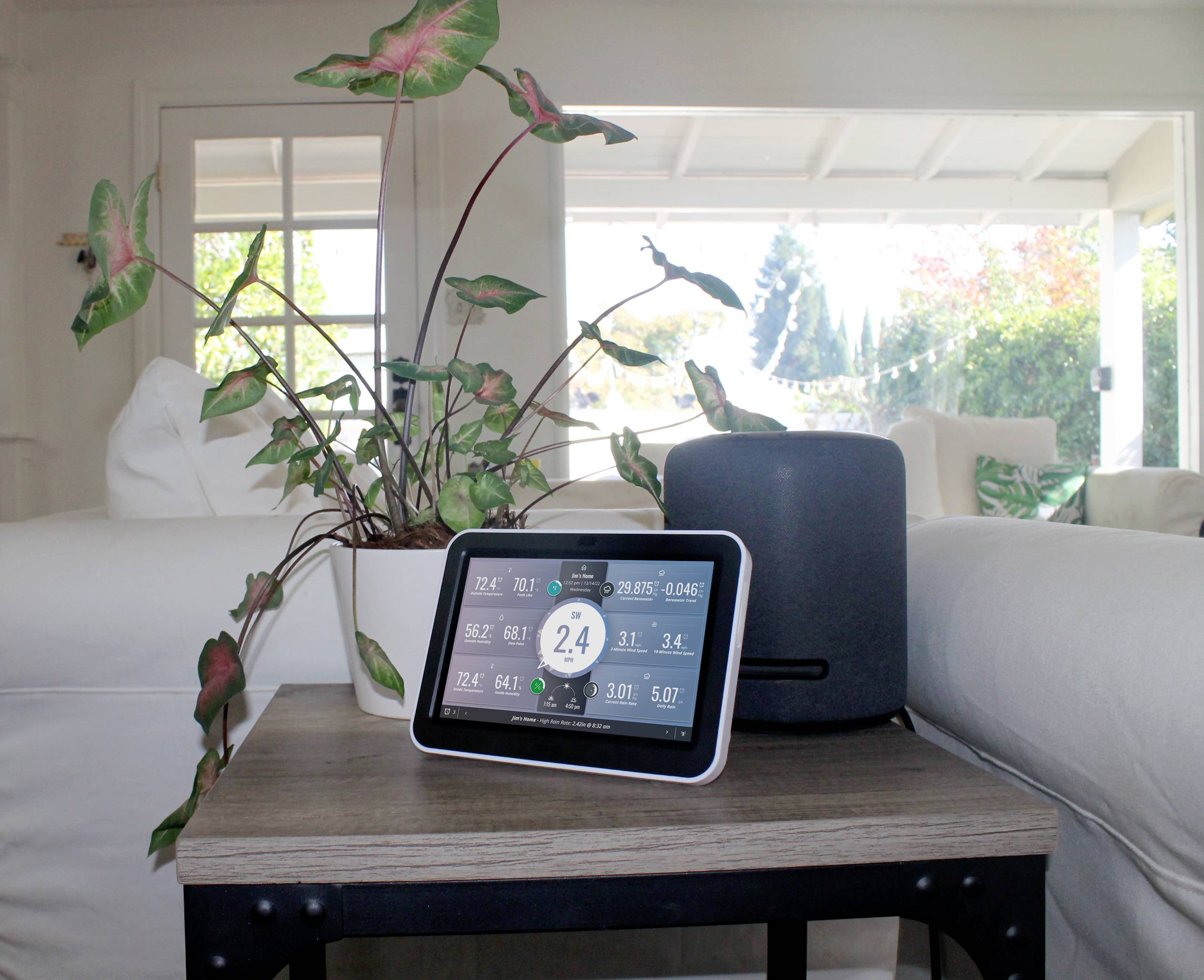 Ein smarter Bildschirm zeigt Wetter- und Raumklimadaten. Daneben steht ein Lautsprecher. Im Hintergrund ein gemütliches Wohnzimmer.