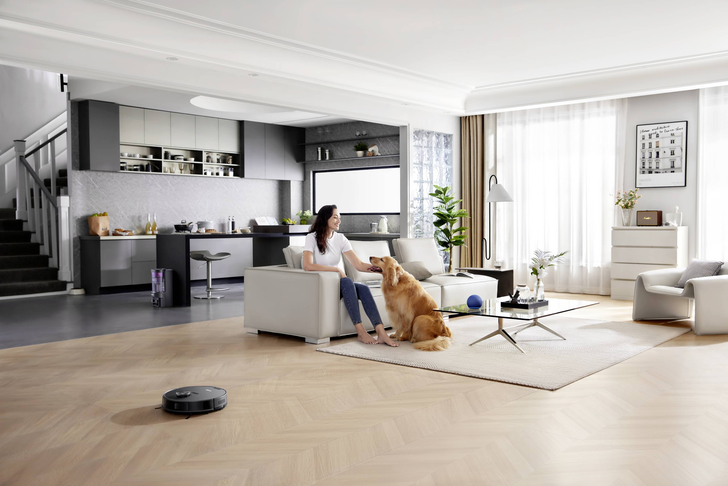 Eine Frau sitzt im Wohnzimmer auf einem Sofa und streichelt einen großen, goldenen Hund. Der Raum ist modern eingerichtet mit einer offenen Küche im Hintergrund.