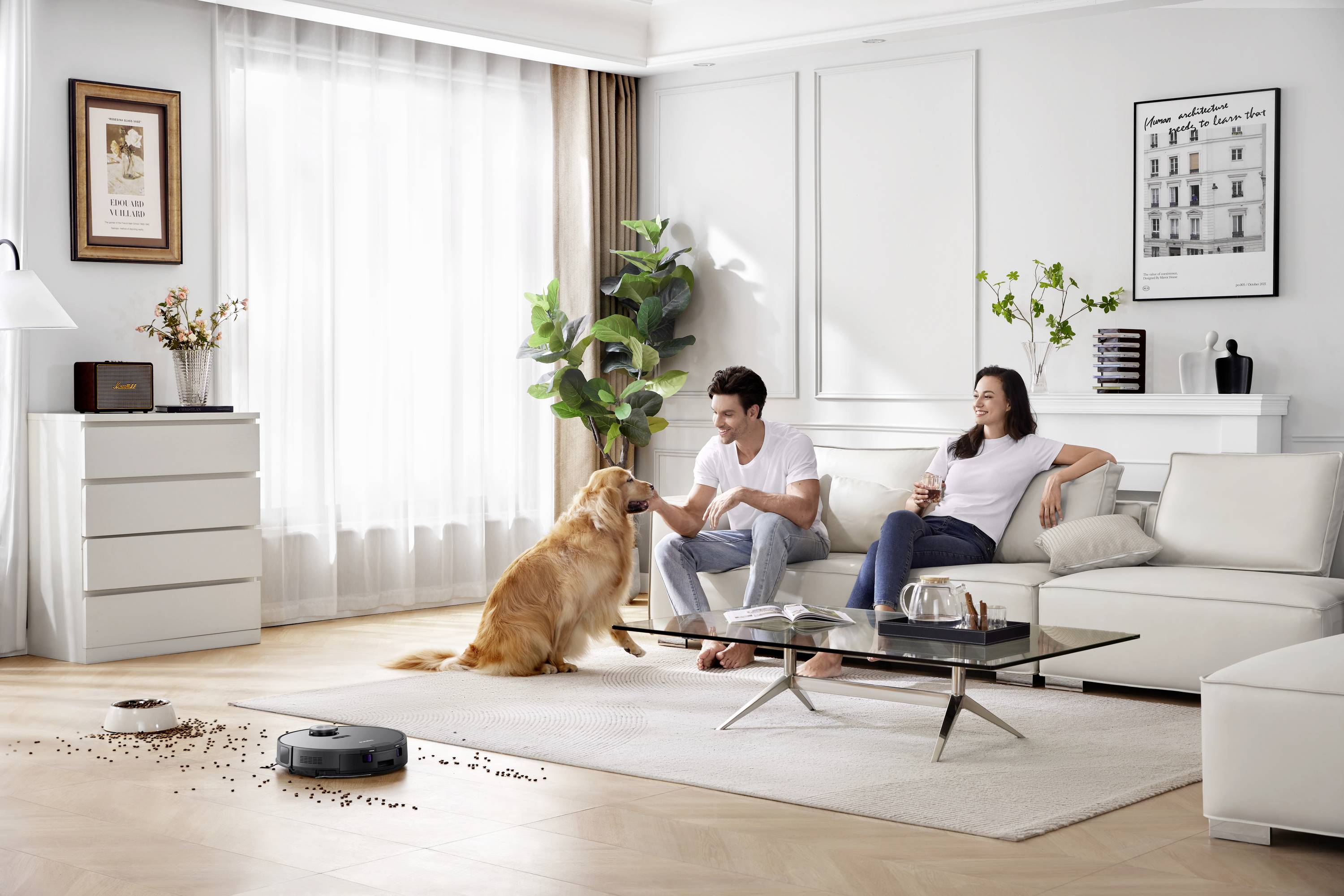 Ein Paar sitzt auf einem Sofa in einem modernen Wohnzimmer, während ein Hund ihnen gegenüber sitzt. Ein Roboterstaubsauger ist aktiv.