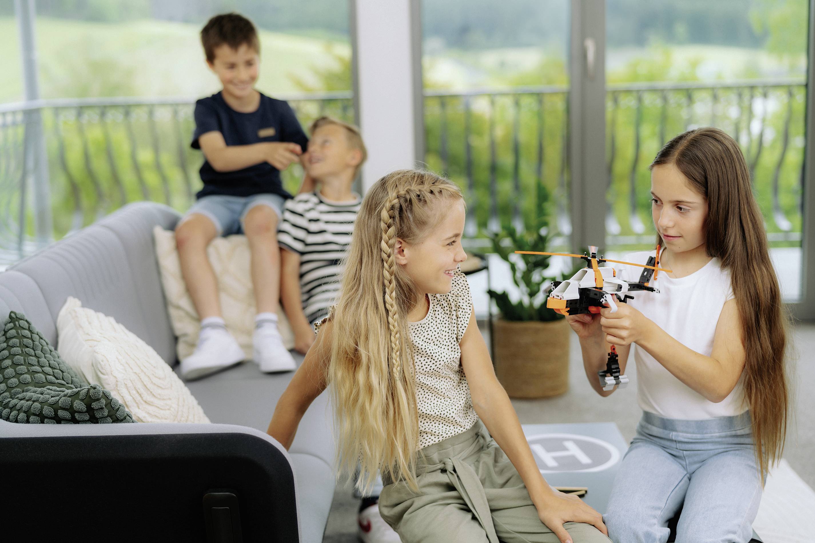 Ein Mädchen zeigt einem anderen Mädchen einen Spielzeughubschrauber im Wohnzimmer. Im Hintergrund spielen zwei Jungen zusammen.