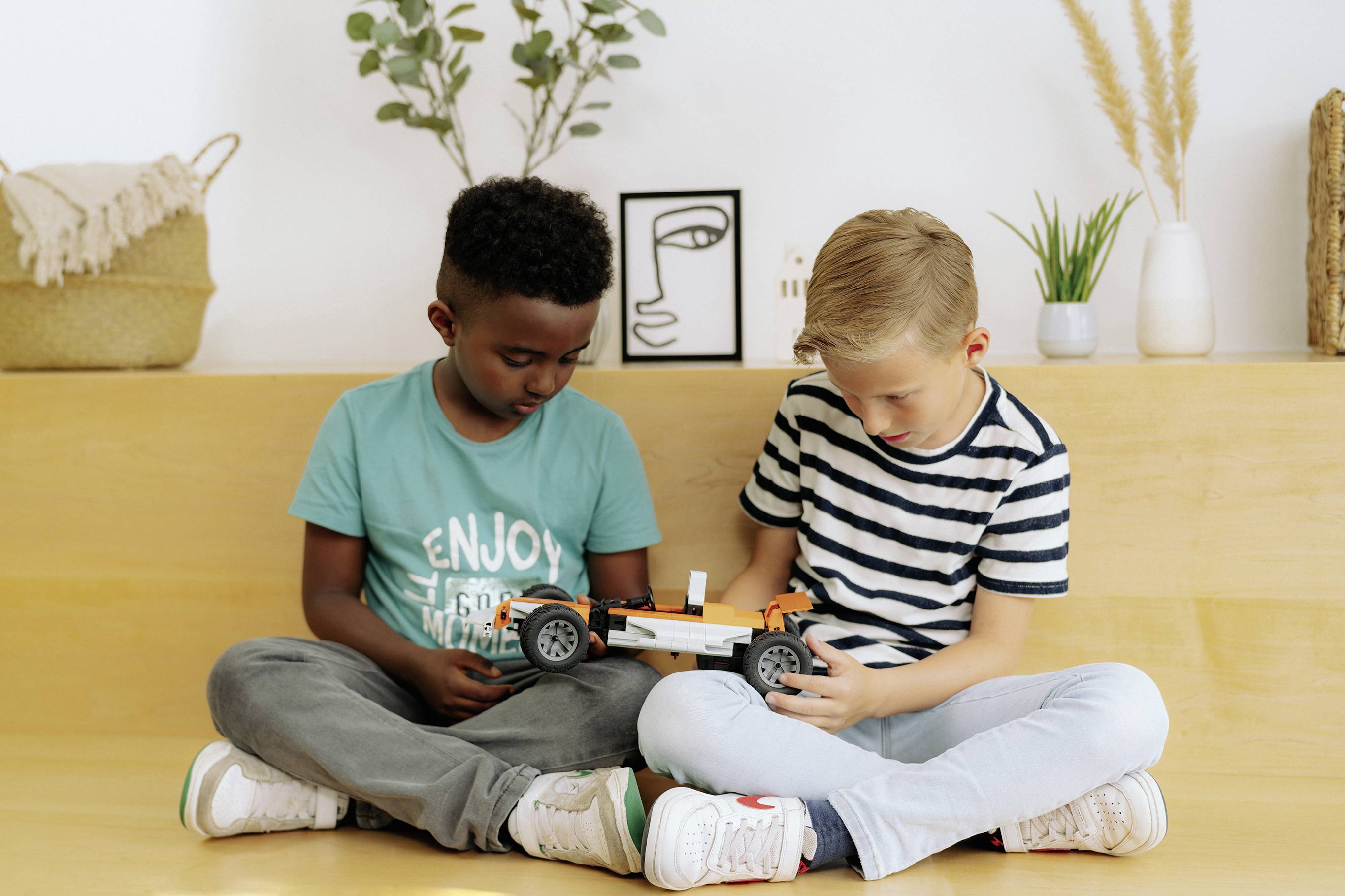 Zwei Kinder sitzen zusammen und untersuchen ein Modellauto. Im Hintergrund sind Pflanzen und dekorative Gegenstände zu sehen.