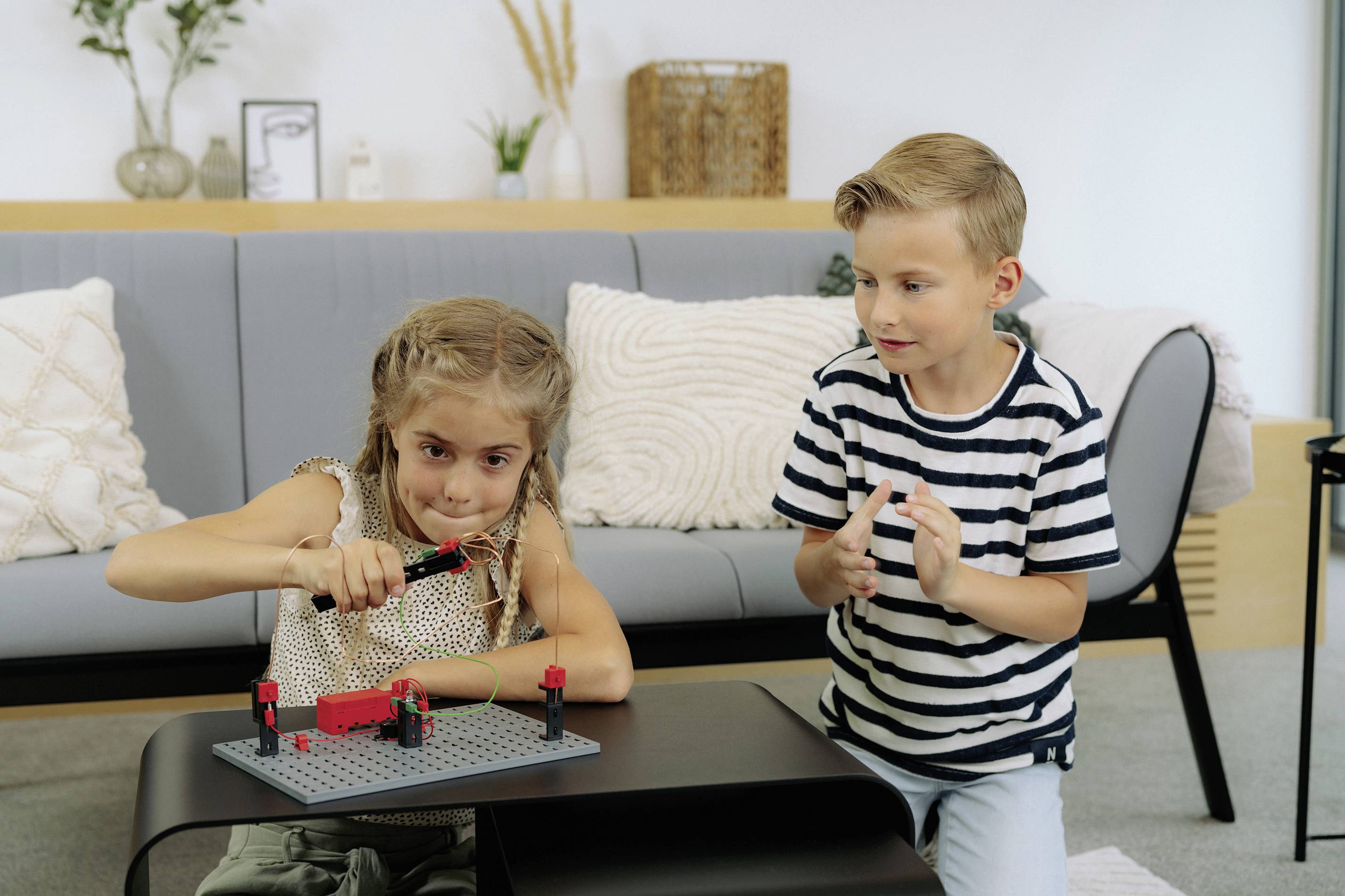 Ein Mädchen und ein Junge spielen konzentriert mit einem Konstruktionsspielzeug auf einem Tisch vor einem Sofa in einem Wohnzimmer.