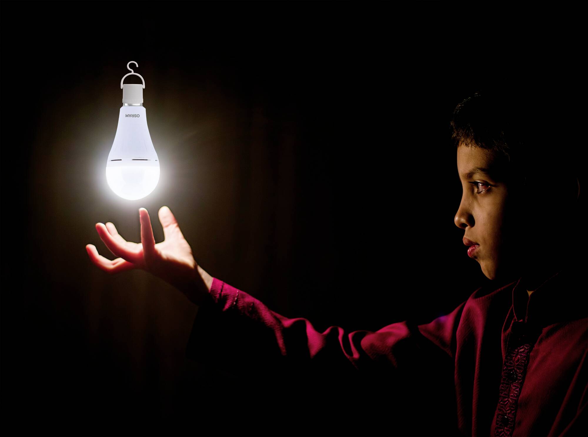 Ein Junge in rotem Hemd schaut konzentriert auf eine schwebende, leuchtende Glühbirne in einer dunklen Umgebung.