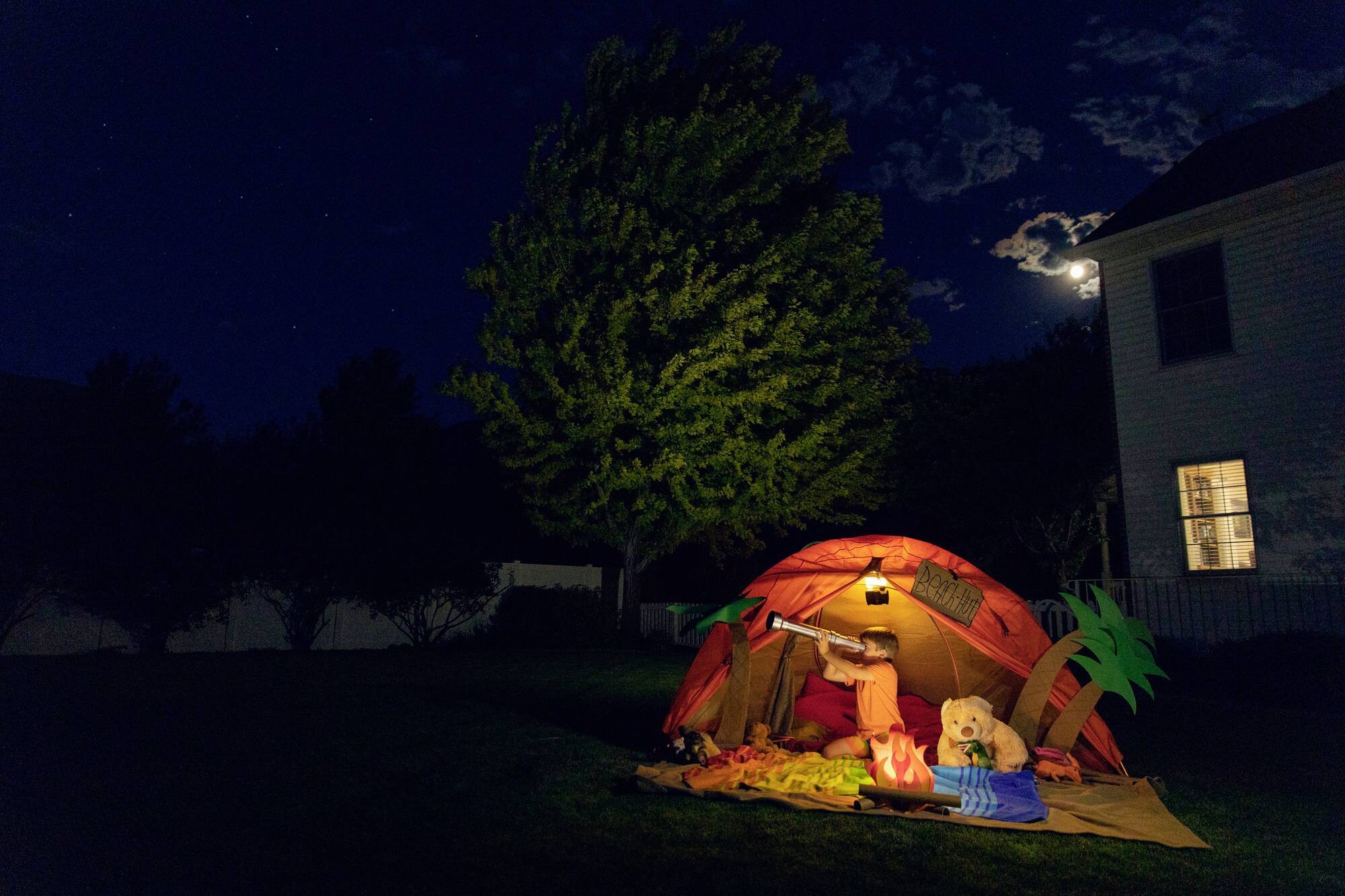 'Kind schaut durch Fernglas im beleuchteten Zelt unter einem Baum bei Nacht, mit Mond im Hintergrund.'