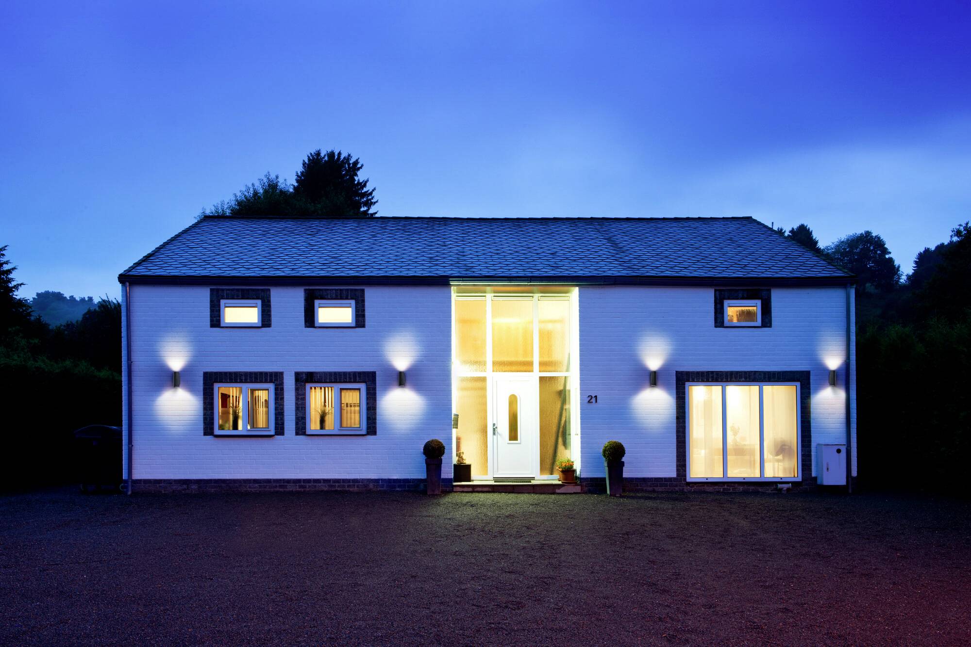 Modernes, beleuchtetes Haus am Abend mit großen Fenstern und Türen, umgeben von Bäumen, vor einem dunkelblauen Himmel.