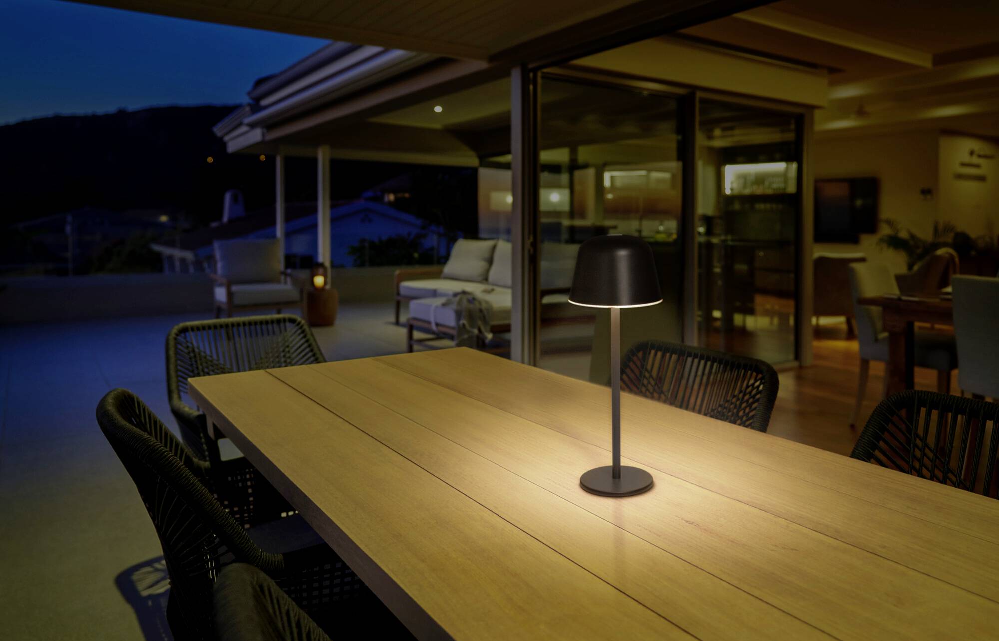 Terrasse bei Nacht mit Holztisch und Lampe. Im Hintergrund beleuchtete Innenräume und Sofa auf der Terrasse.