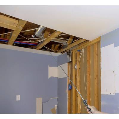 Person zeigt mit einem Zeigestock auf freiliegende Rohre und Kabel in einer geöffneten Decke eines Teils eines renovierungsbedürftigen Zimmers.