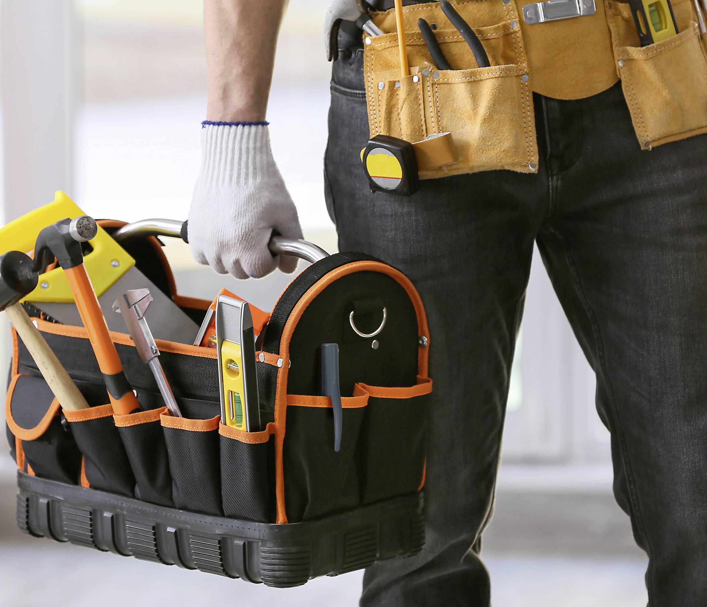 Eine Person hält eine Werkzeugtasche mit Hämmern, Schraubendrehern und anderen Werkzeugen. Sie trägt einen Werkzeuggürtel.