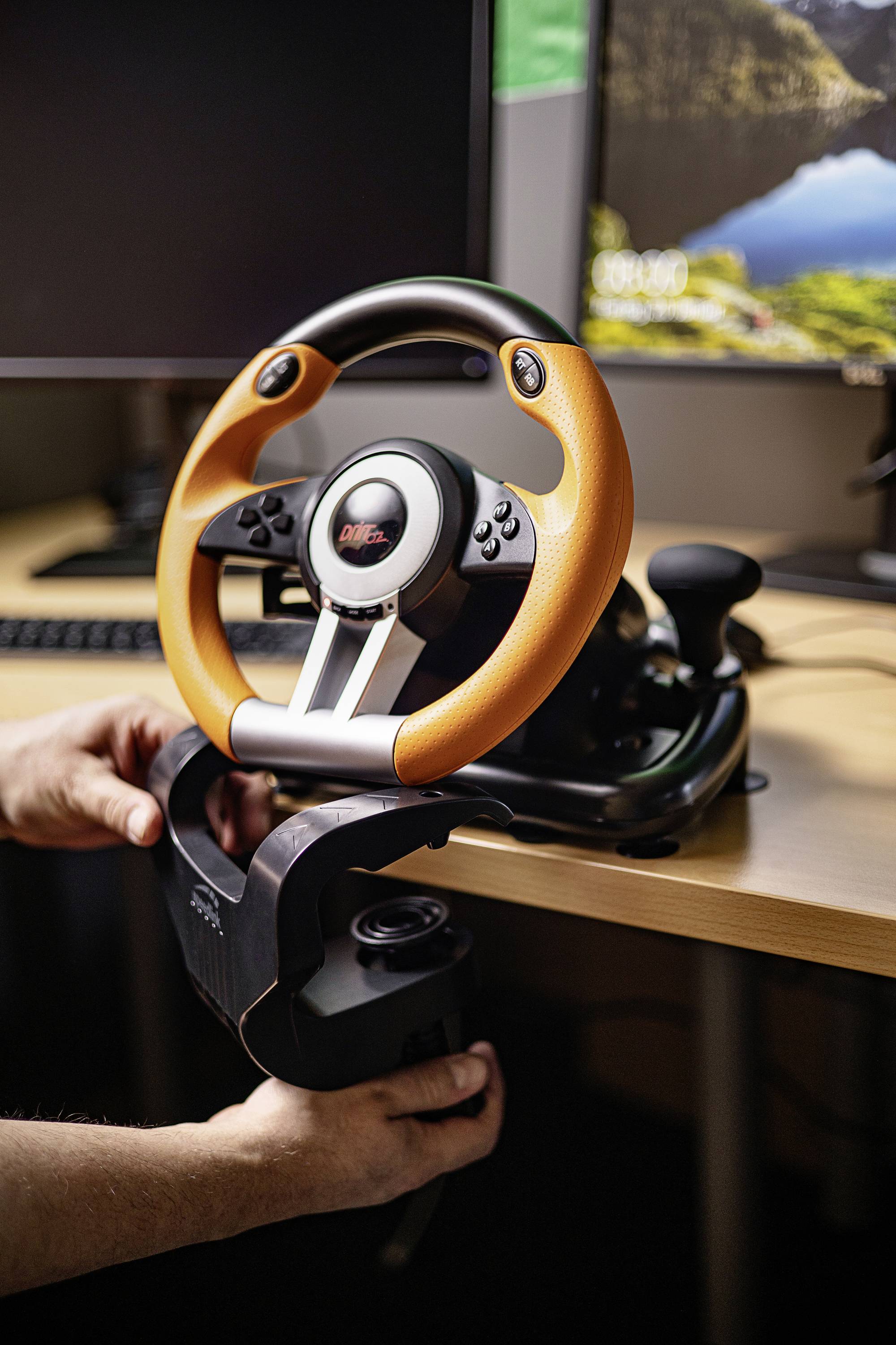 Eine Person befestigt ein orange-schwarzes Lenkrad an einem Schreibtisch. Im Hintergrund sind ein Monitor und eine Tastatur sichtbar.