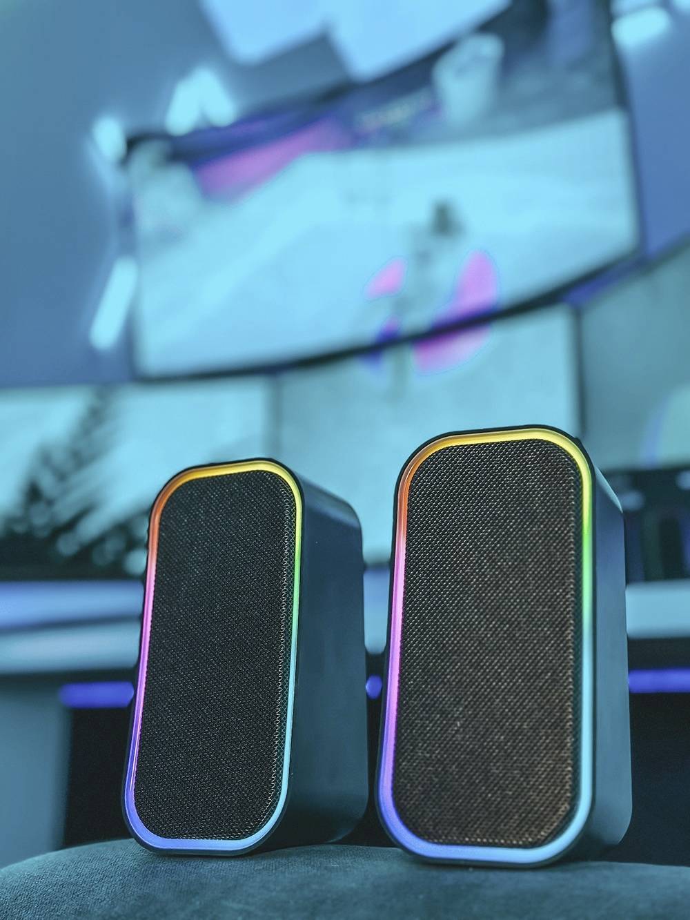 Zwei Lautsprecher mit Regenbogenlichtern vor einem Hintergrund mit gebogenen Bildschirmen, die ein Videospiel darstellen.