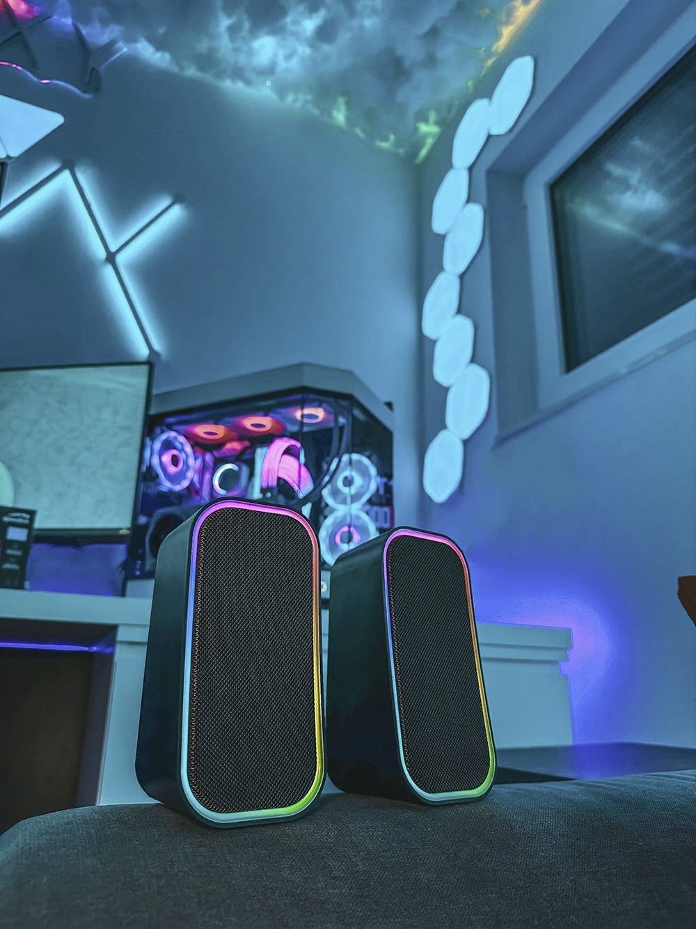 Zwei dunkle Lautsprecher mit bunten LED-Leuchten vor einem Gaming-PC und beleuchtetem Raumhintergrund.