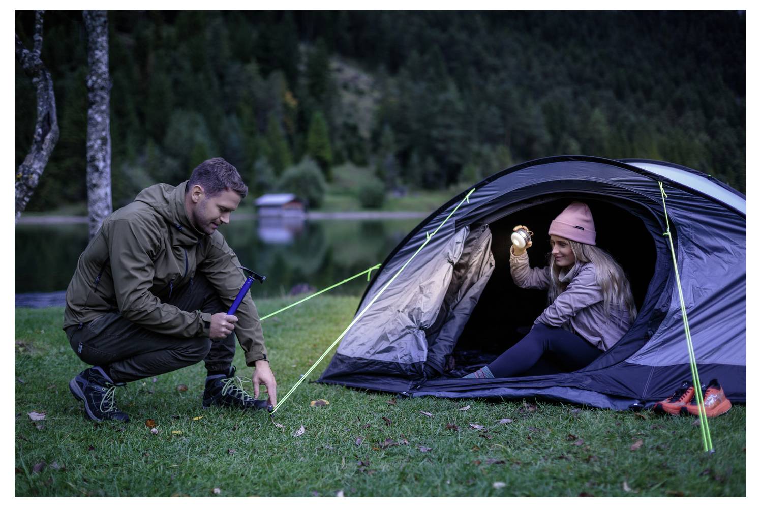 Ein Mann kniet neben einem Campingzelt, während eine Frau darin lächelt und eine Taschenlampe bei Einbruch der Dunkelheit neben einem See hält.