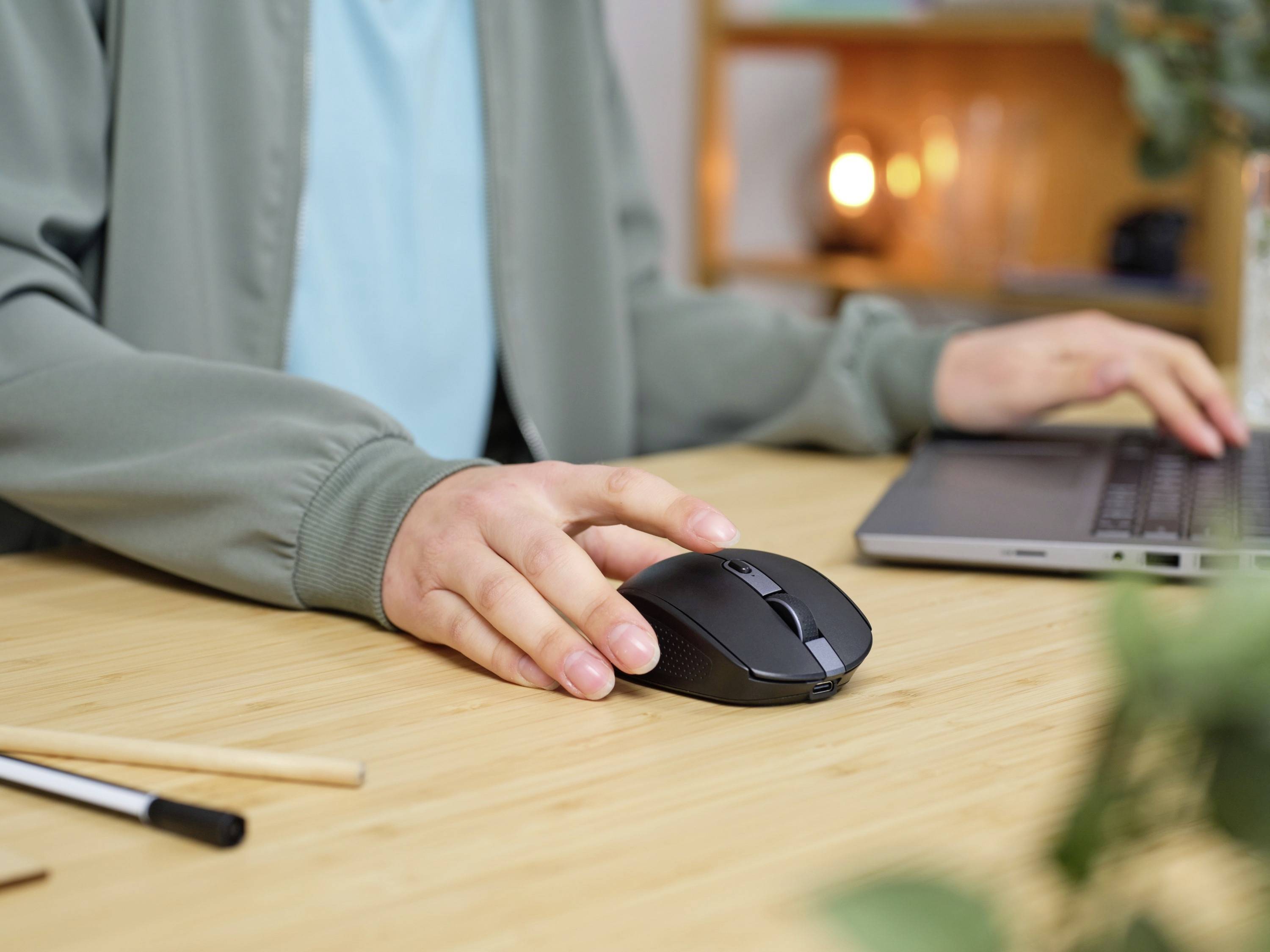 Eine Person arbeitet am Laptop, verwendet eine kabellose Maus, sitzt an einem Tisch mit Stiften im Vordergrund; Hintergrund zeigt Regale.