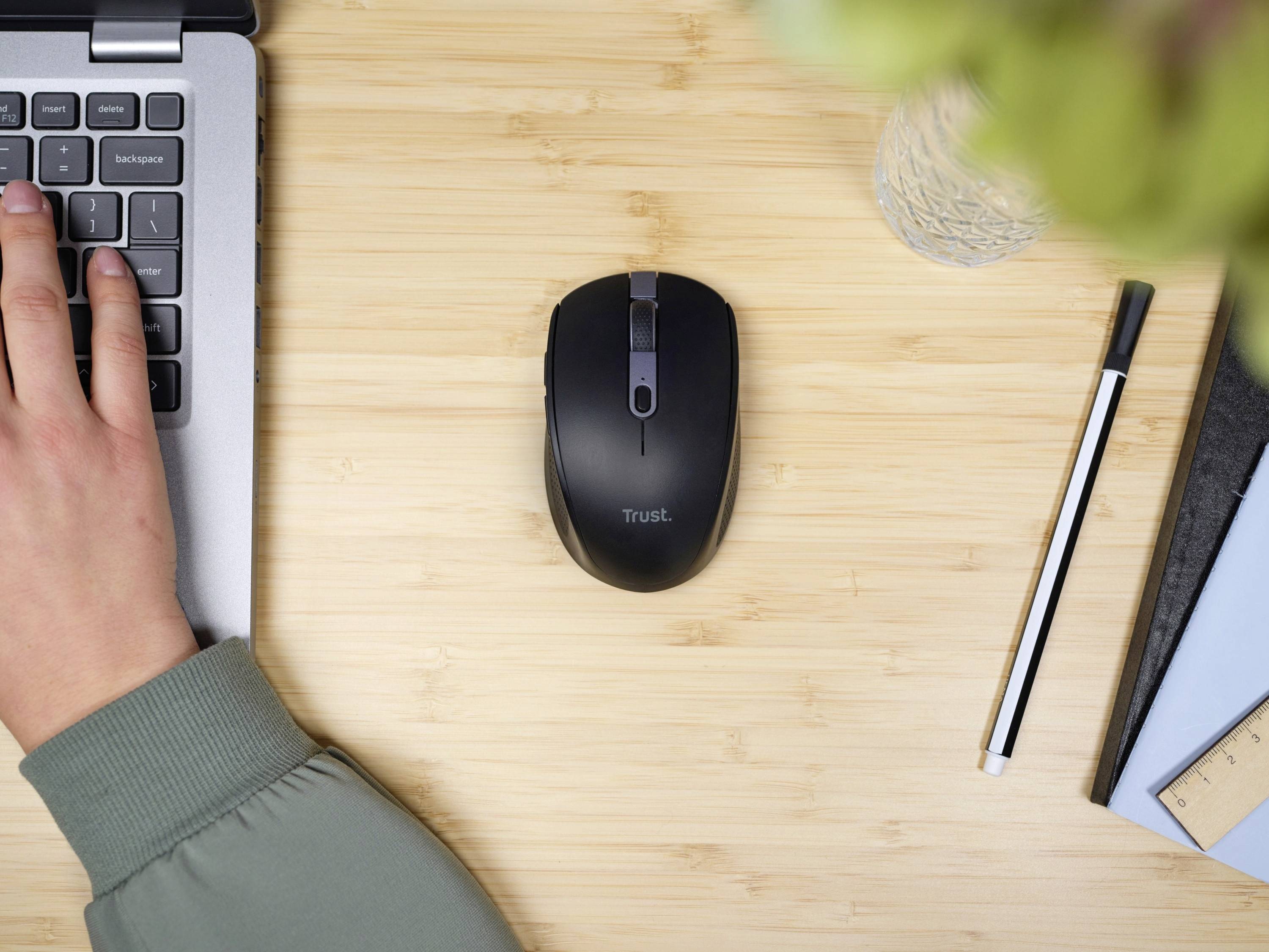 Ein Laptop mit einer Hand auf der Tastatur, daneben eine schwarze kabellose Maus auf einem Holztisch, daneben ein Stift und ein Notizbuch.