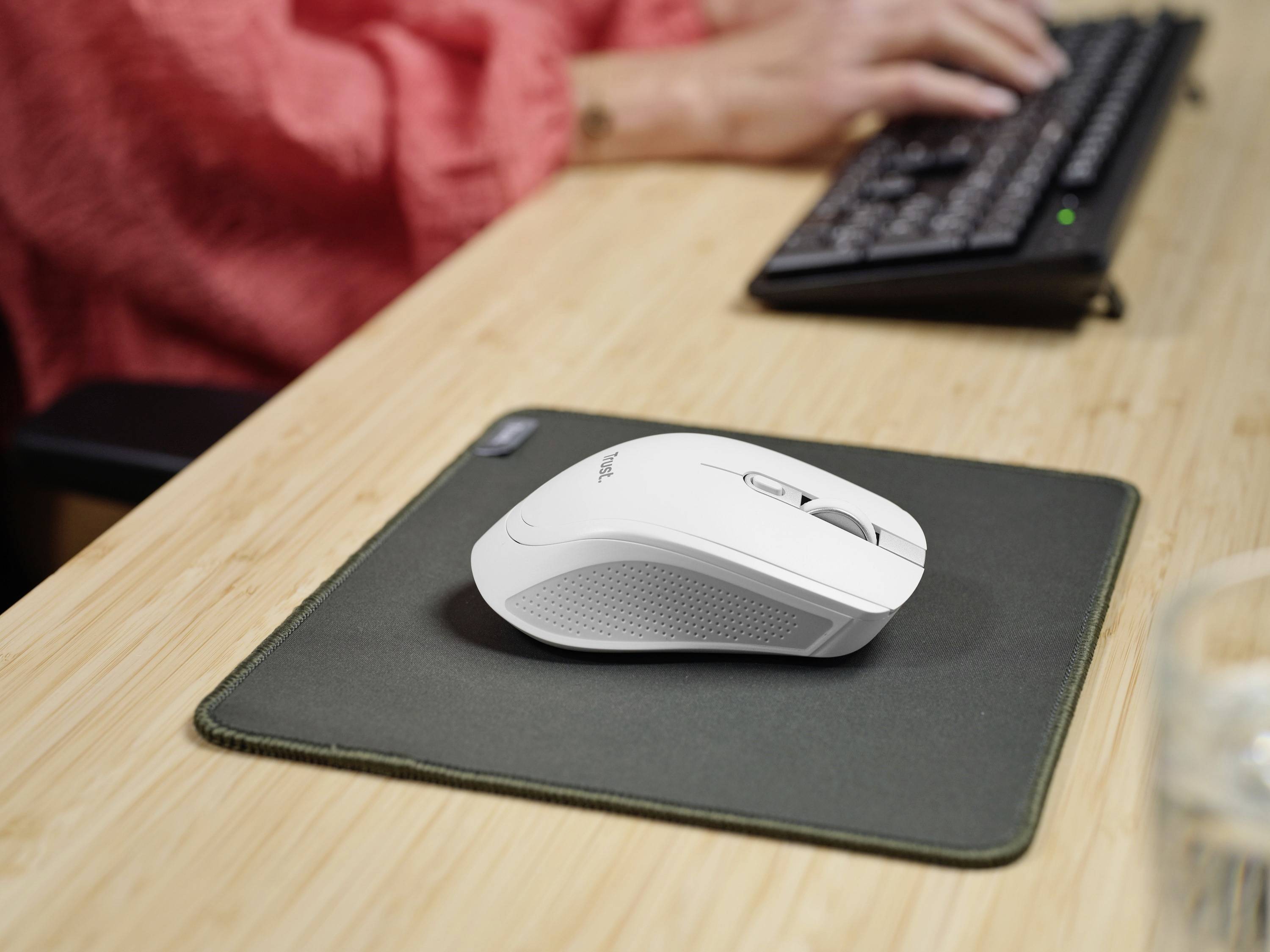 'Eine weiße Computermaus auf einem grünen Mauspad, eine Person tippt auf einer Tastatur im Hintergrund.'