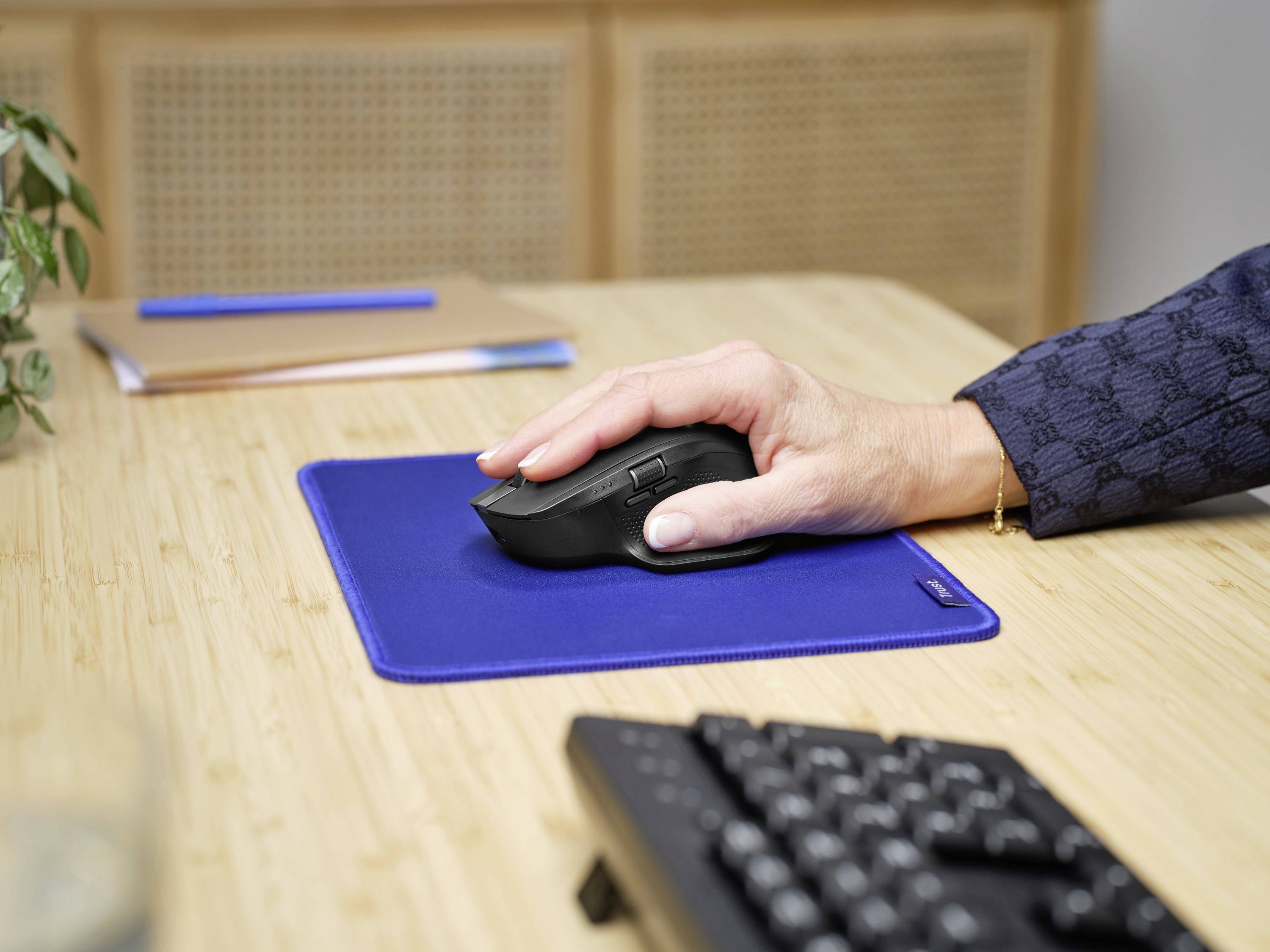 Eine Hand bedient eine schwarze Computermaus auf einem blauen Mauspad, das auf einem Holztisch liegt. Im Hintergrund ein Computerkeyboard.