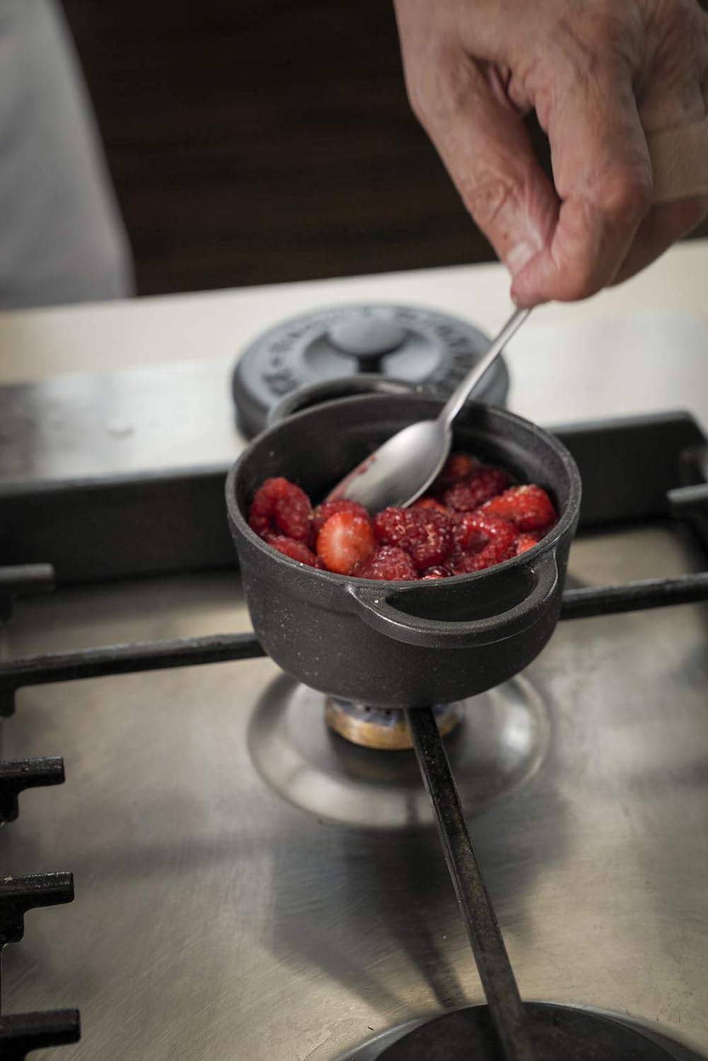 Eine Hand hält einen Löffel über einem kleinen Topf auf einem Gasherd. Im Topf sind Erdbeeren und Himbeeren.