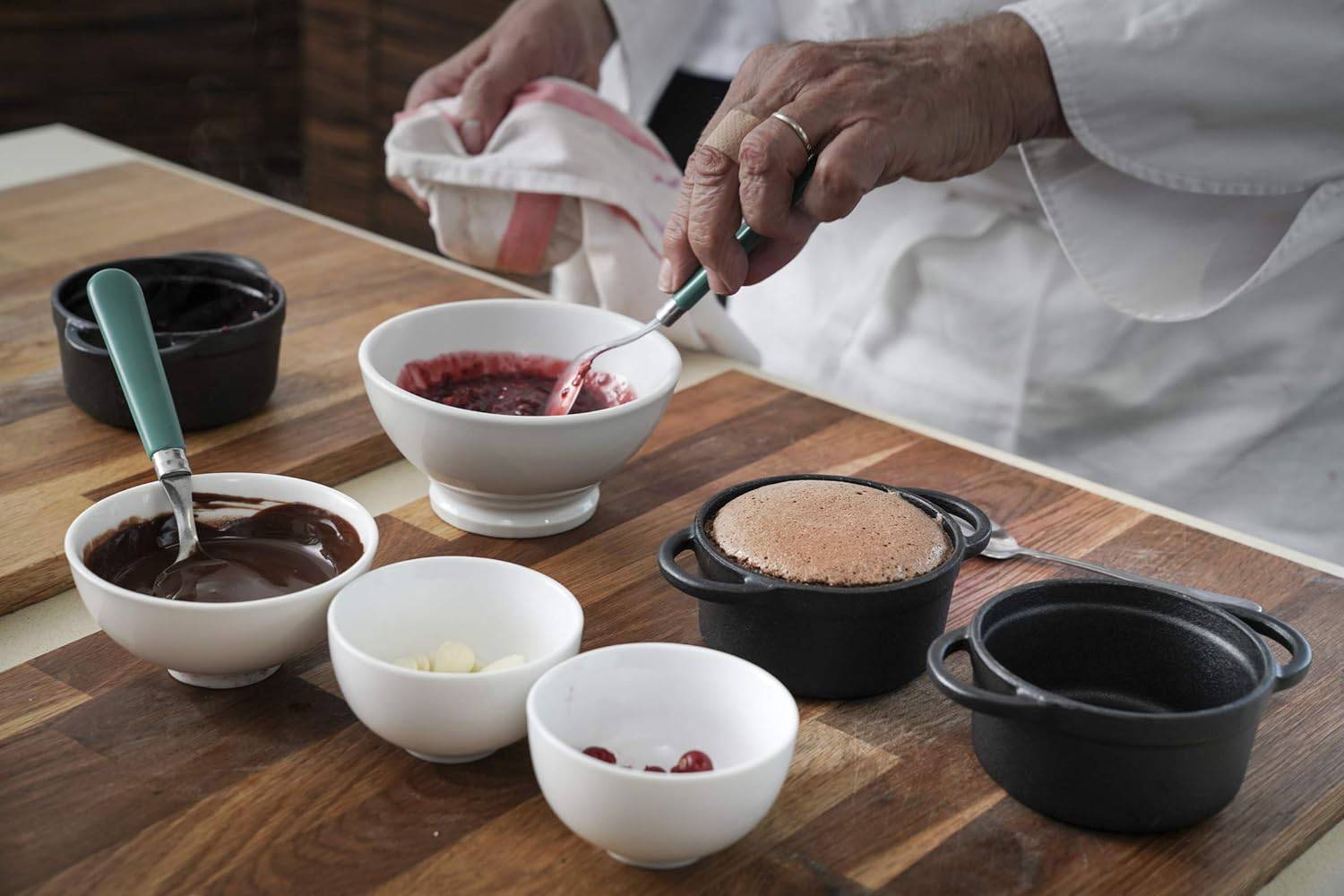 Ein Koch bereitet ein Dessert zu. Auf einem Tisch stehen Schalen mit Schokolade, roten Beeren und ein kleiner Kuchen in einem Töpfchen.