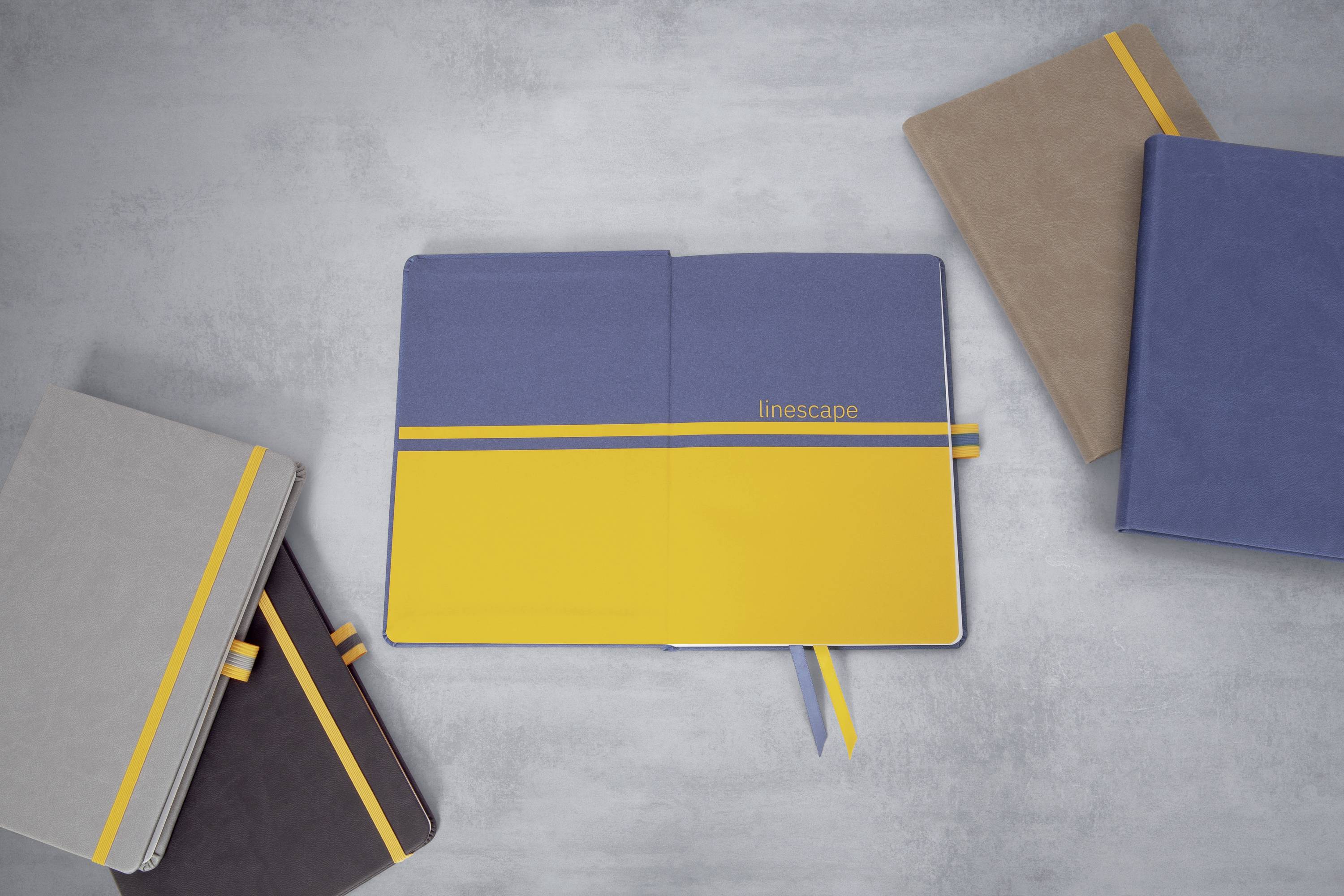 Ein blaues und gelbes Notizbuch mit der Aufschrift 'linescape' liegt geöffnet auf einem grauen Tisch. Mehrere geschlossene Notizbücher liegen daneben.