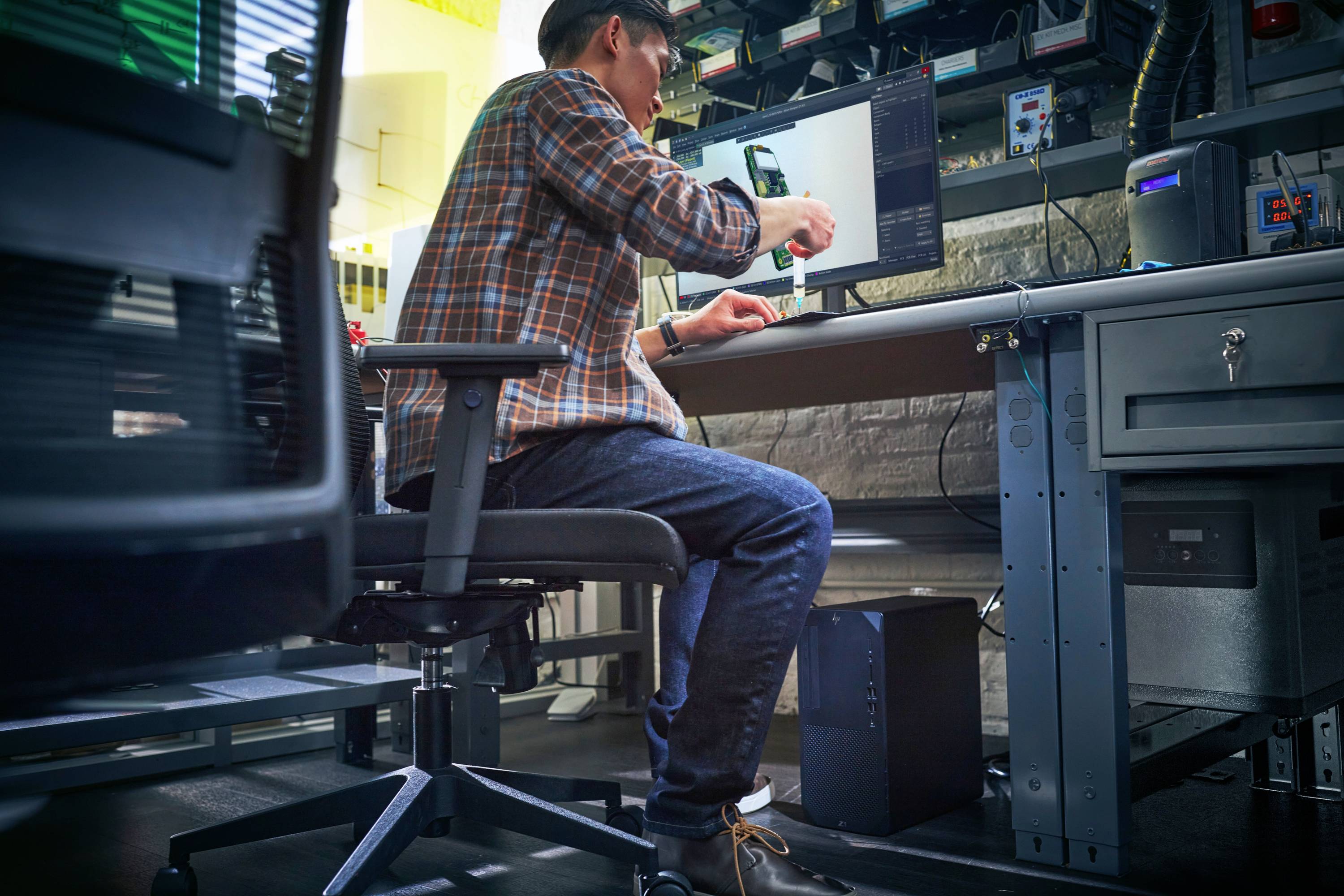 Eine Person sitzt an einem Schreibtisch in einer Werkstatt oder einem Labor und arbeitet an einem Computer.