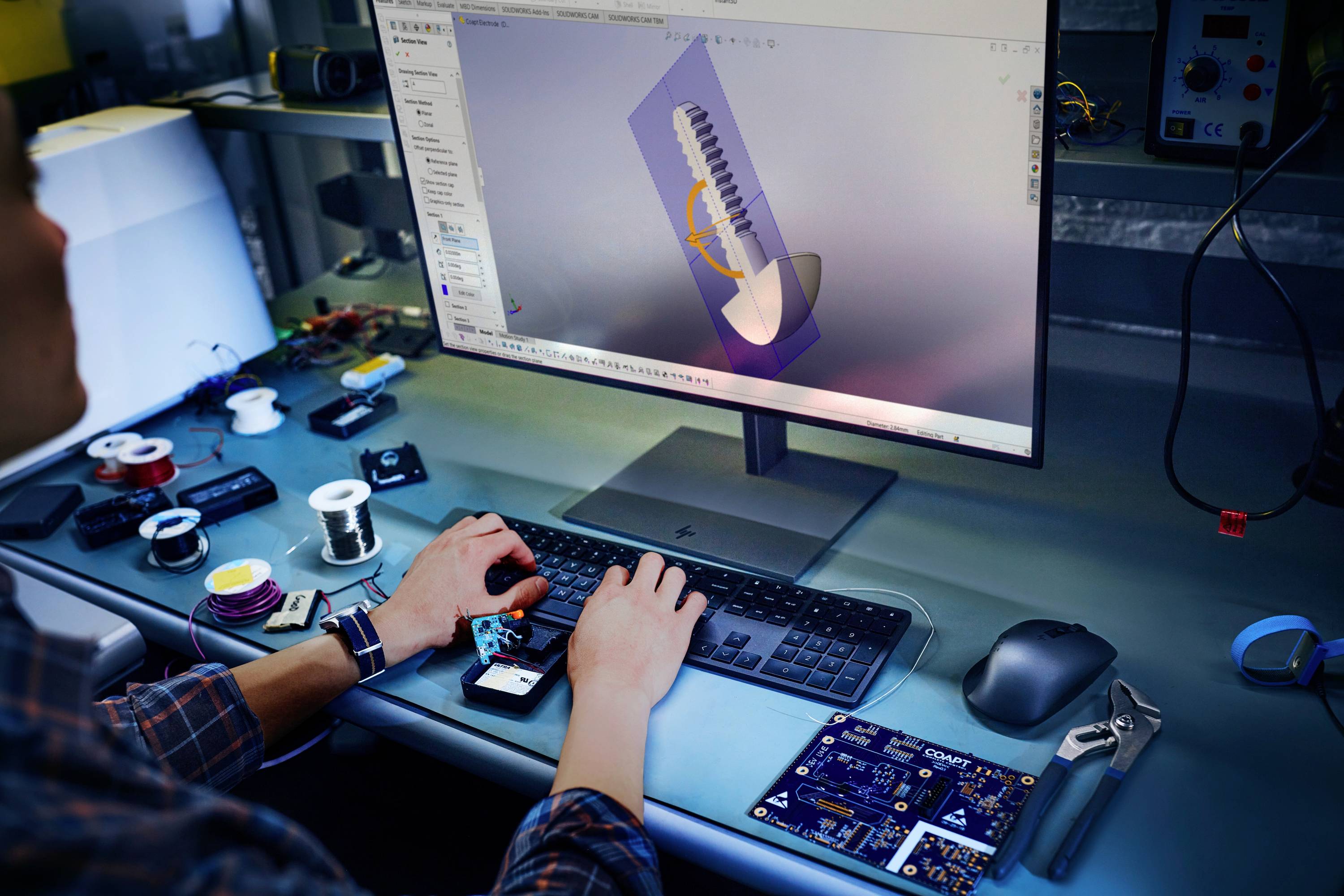 Eine Person arbeitet an einem Computer mit einem 3D-Design-Programm, um ein technisches Bauteil zu modellieren. Elektronikteile und -werkzeuge sind auf dem Tisch verstreut.