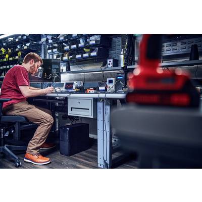 Ein Mann in einem Labor sitzt an einem Tisch und arbeitet an elektronischen Geräten. Der Raum ist mit Regalen voller Ausrüstung ausgestattet.