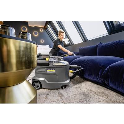 Person reinigt einen blauen Samtsofa mit einem professionellen Teppichreiniger in einem modernen, gut beleuchteten Raum mit schrägen Fenstern.