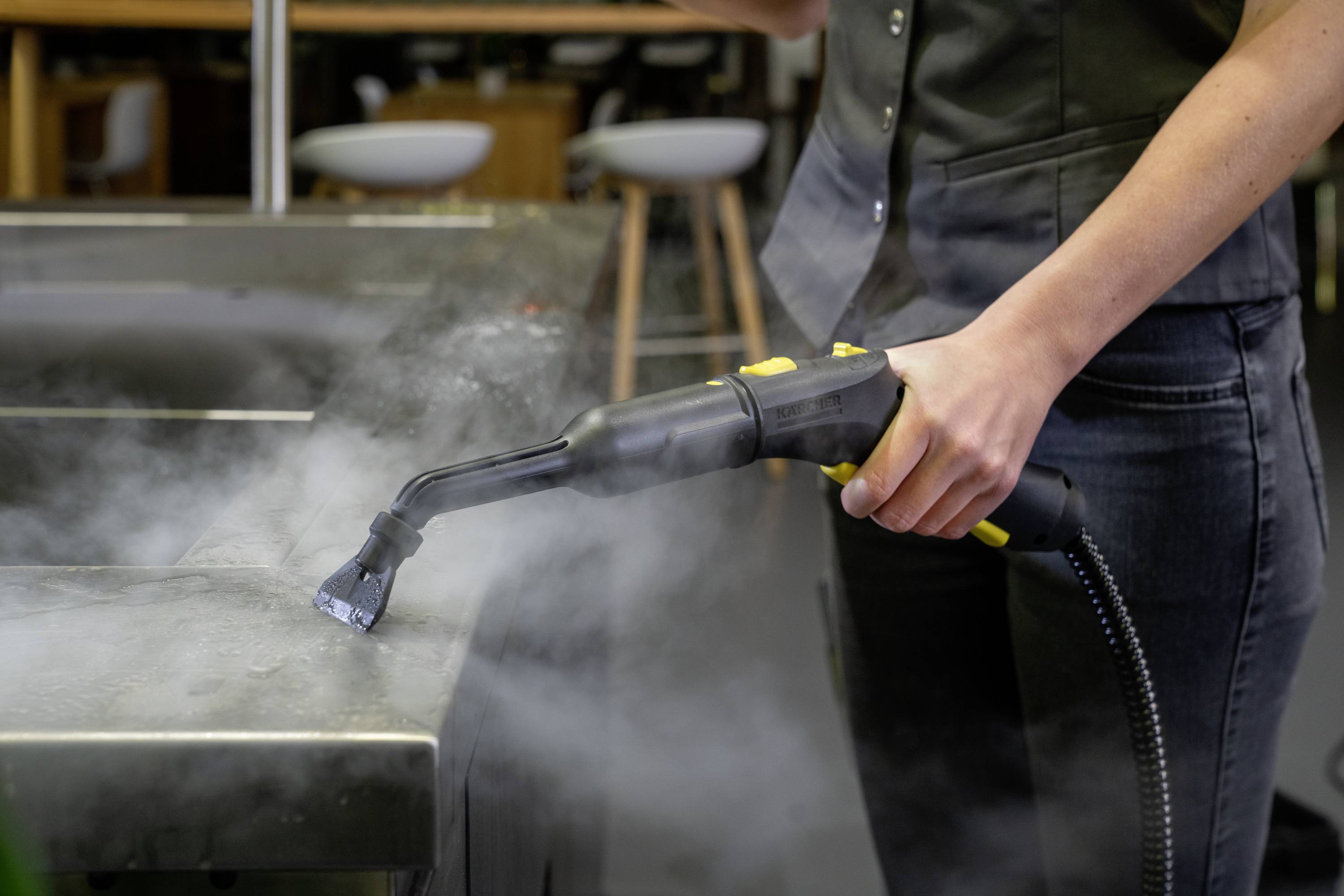 Eine Person dampft eine Metalloberfläche mit einem Dampfreiniger ab. Dampf steigt auf, während die Reinigung durchgeführt wird.